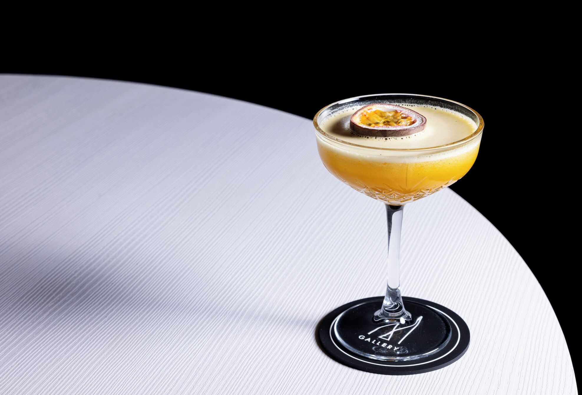 Un cocktail servi dans un verre à martini, posé sur une table blanche avec un dessous de verre noir.