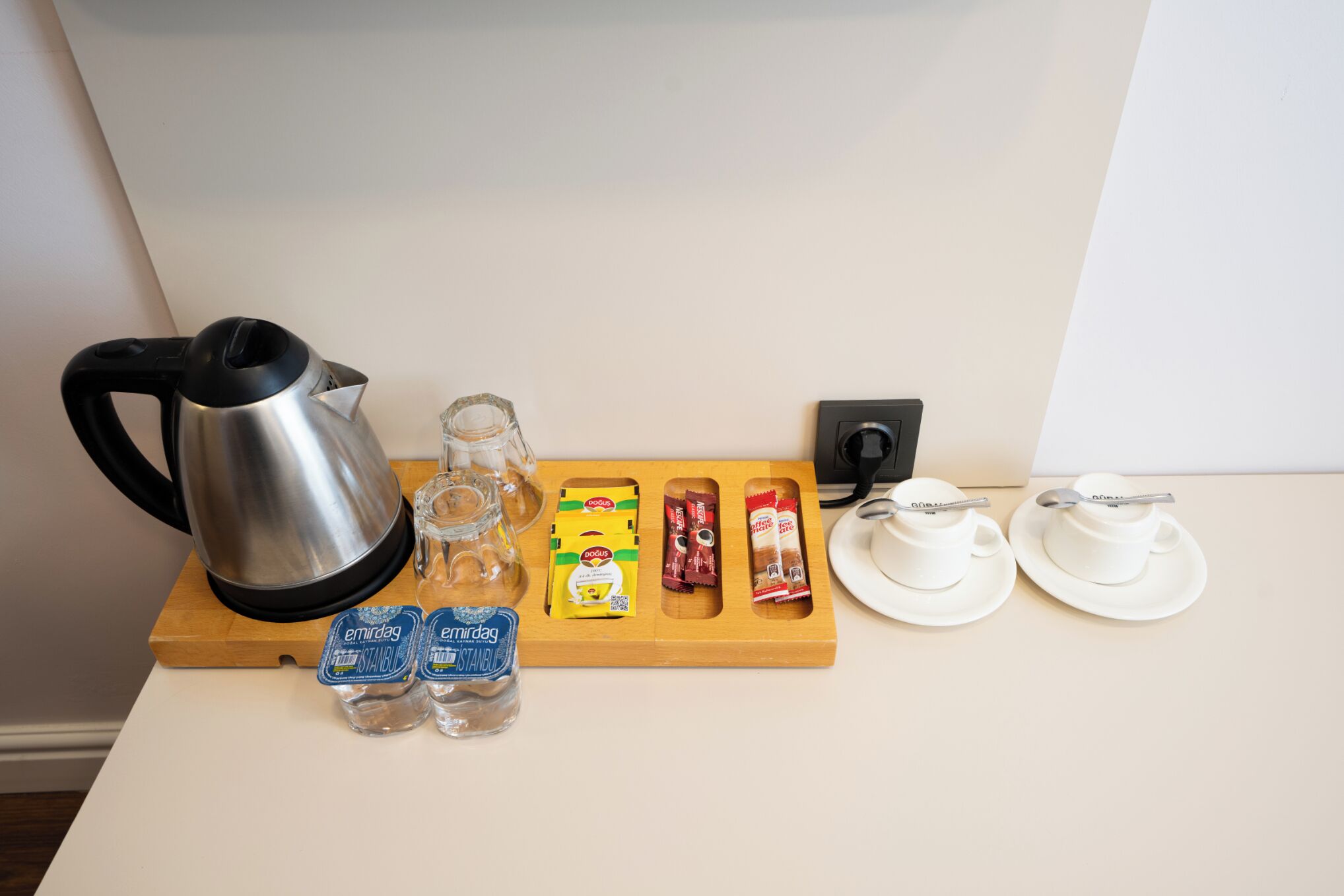 Un coin cuisine comprenant une bouilloire, des bocaux en verre, des en-cas emballés et deux tasses blanches disposées sur un plateau en bois.