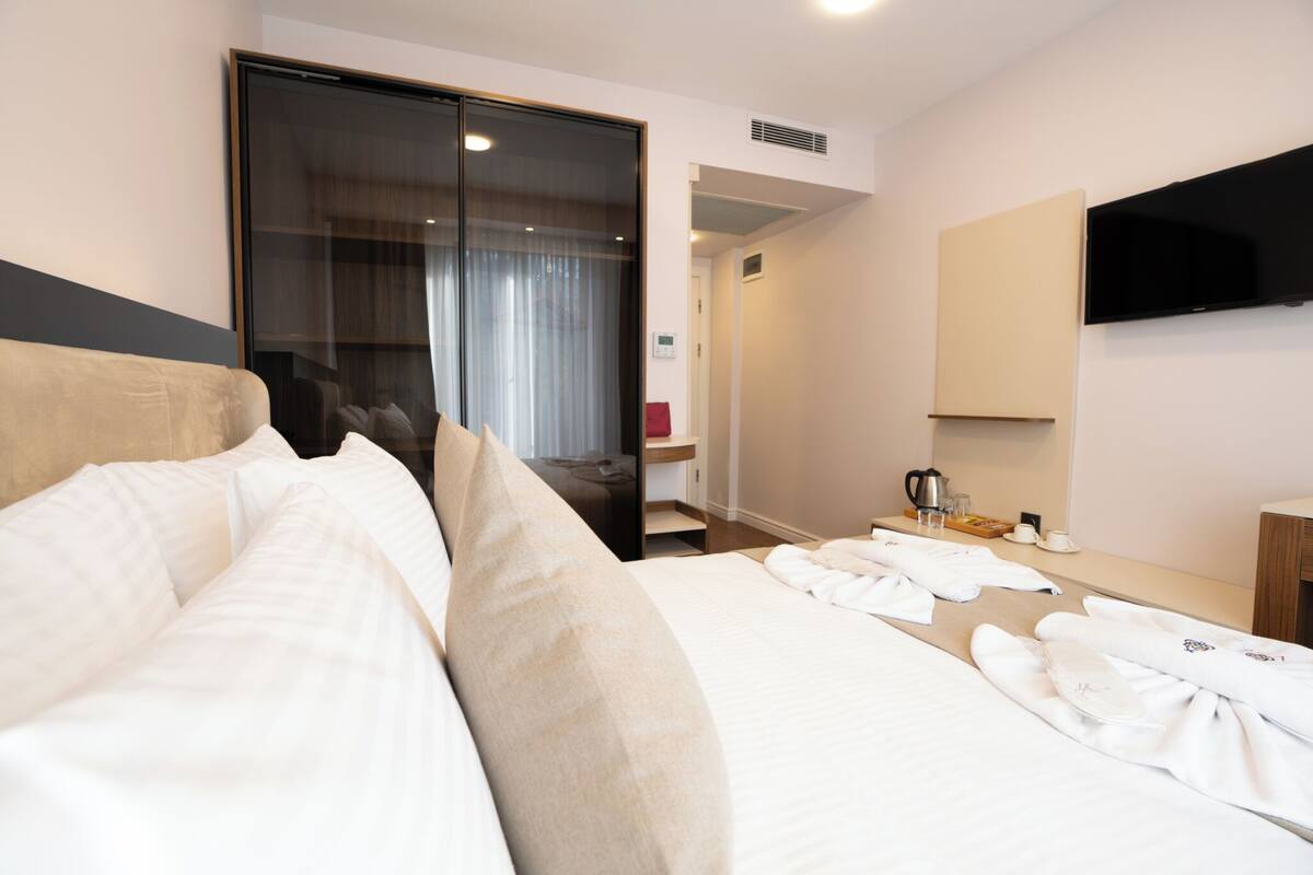 A hotel room with a large bed, a wardrobe, a desk with a coffee maker, and a flat-screen TV mounted on the wall.