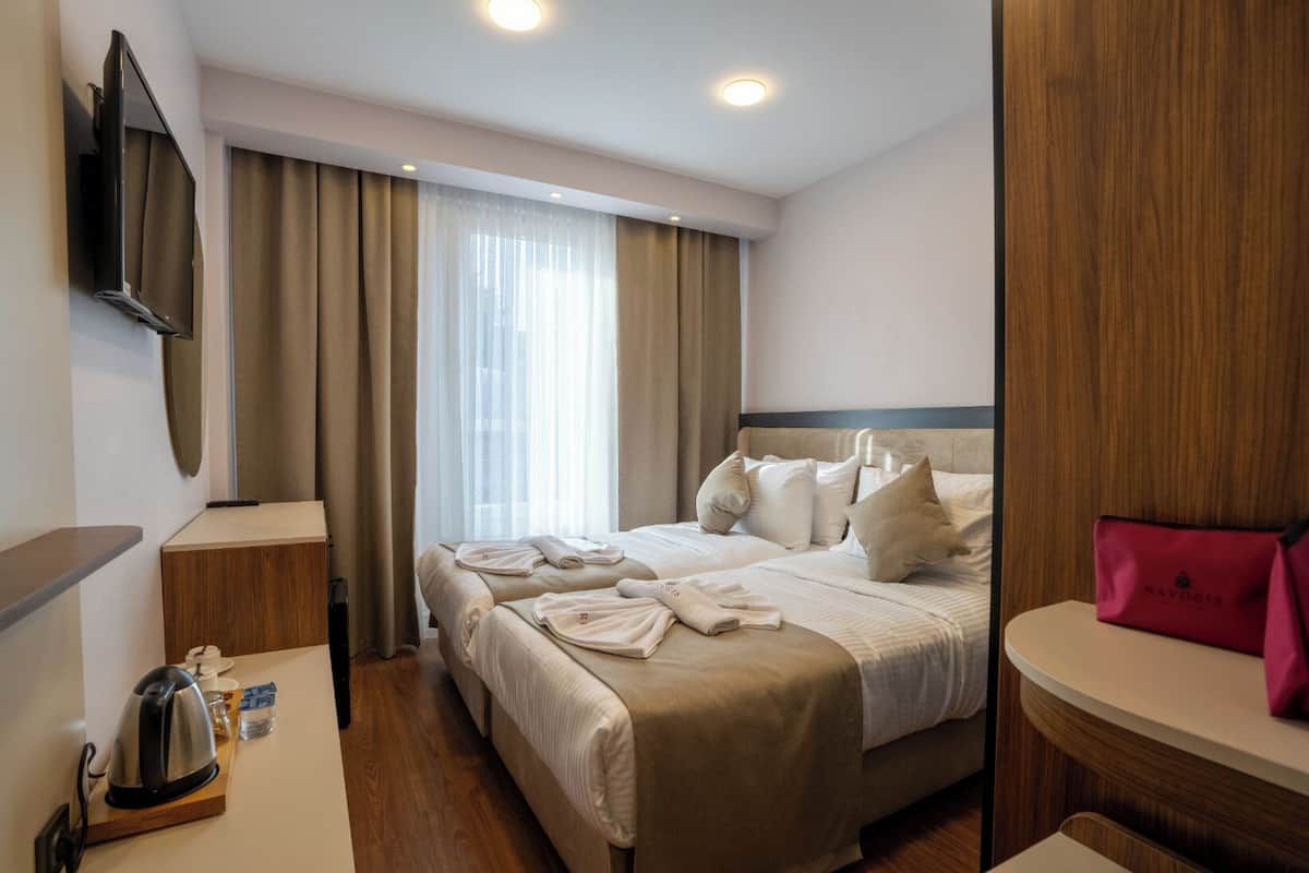 A hotel room with a bed, a TV mounted on the wall, a bedside table with a kettle and a glass of water, and a wooden headboard.