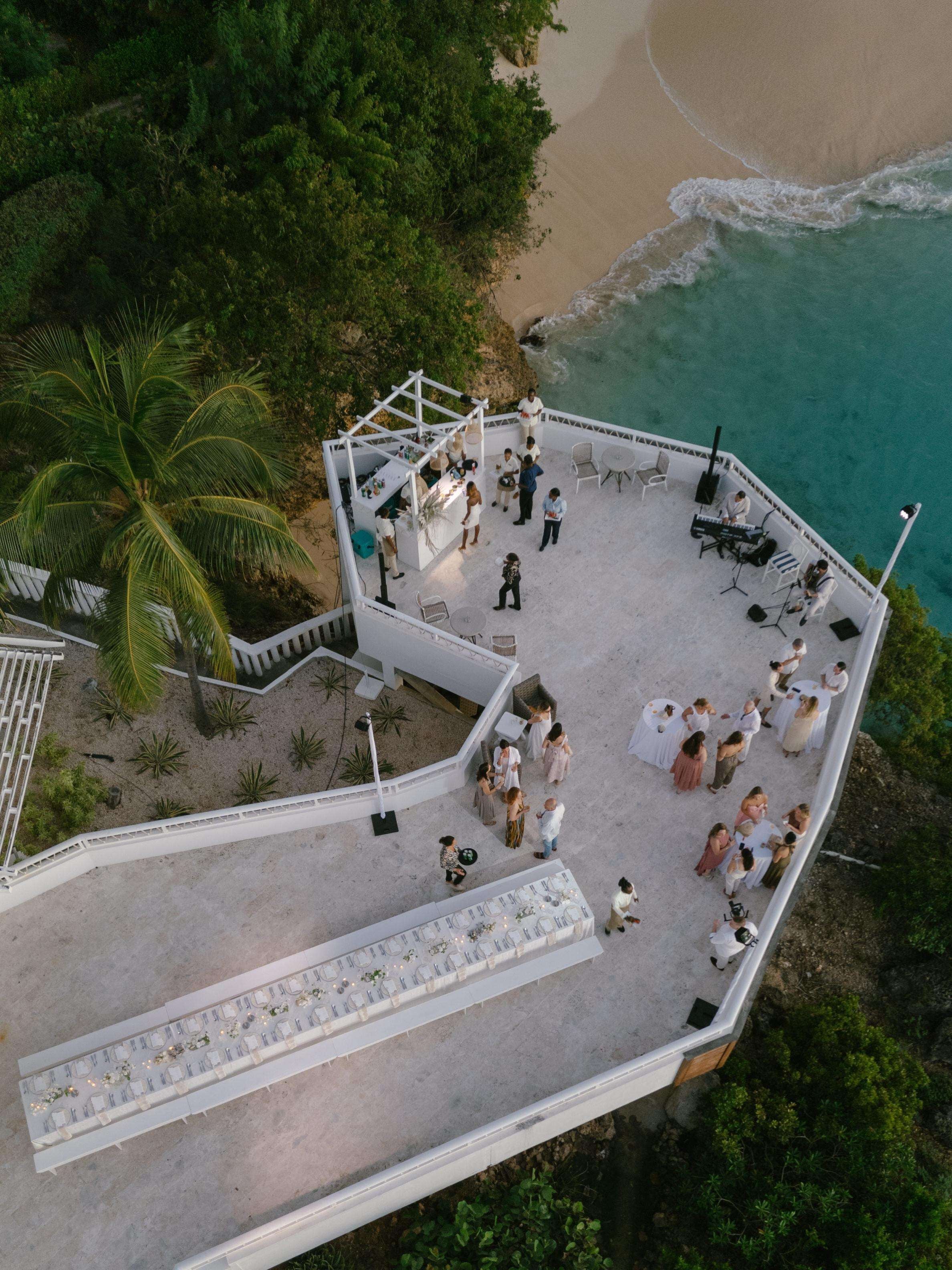An outdoor wedding setup with a white tent, tables, and chairs, overlooking a beach and ocean.
