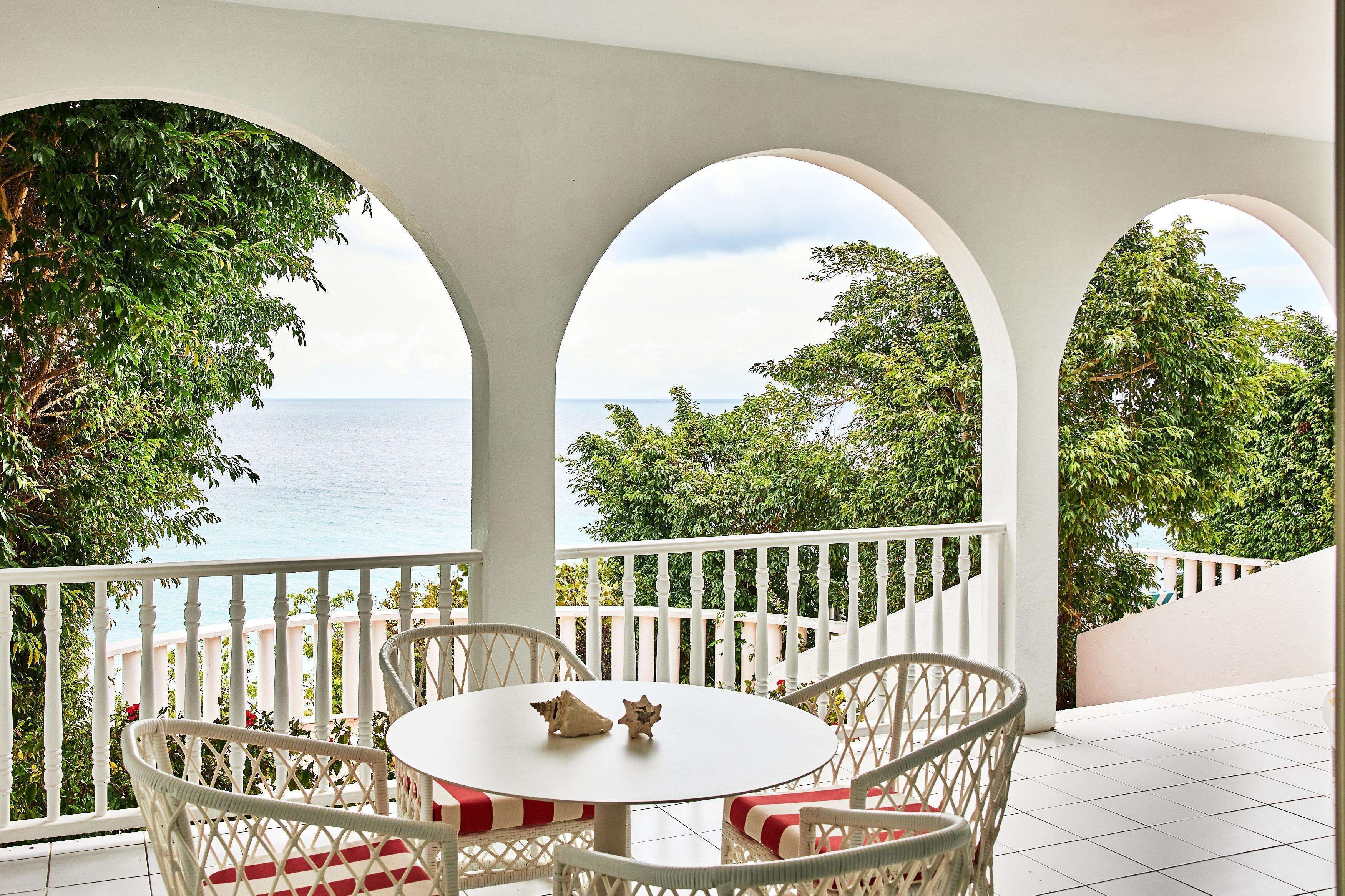 Un balcón con barandilla blanca, una mesa redonda con dos sillas y vistas al mar y la vegetación.