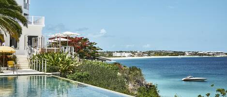 A pool with a view of the sea and a boat, surrounded by greenery and a building with balconies.