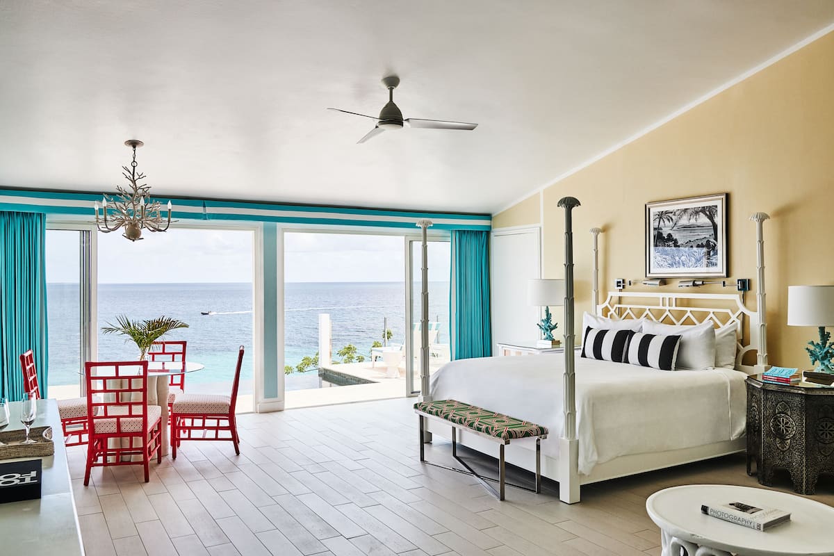 A hotel room with a bed, a ceiling fan, a view of the ocean, a dining area with chairs, and a small table with a book.
