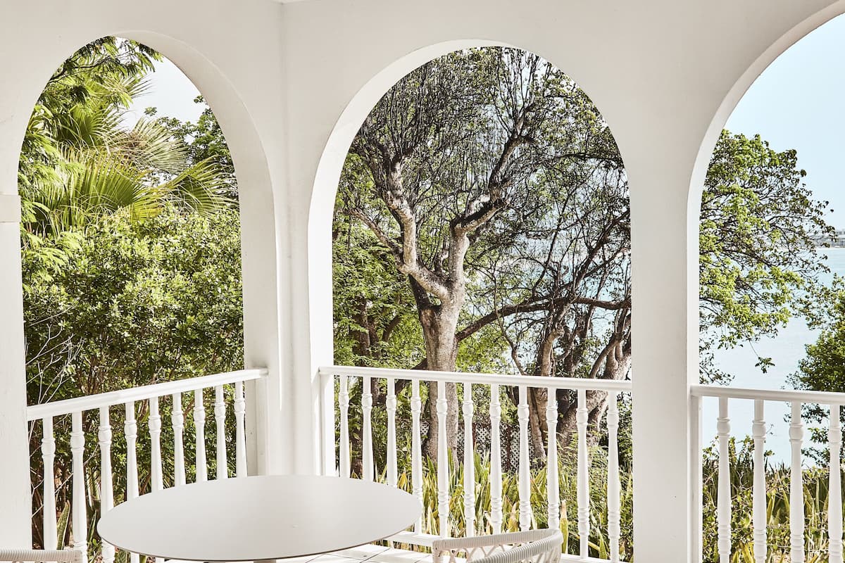 A covered outdoor seating area with white furniture and arches, overlooking a lush garden and trees.