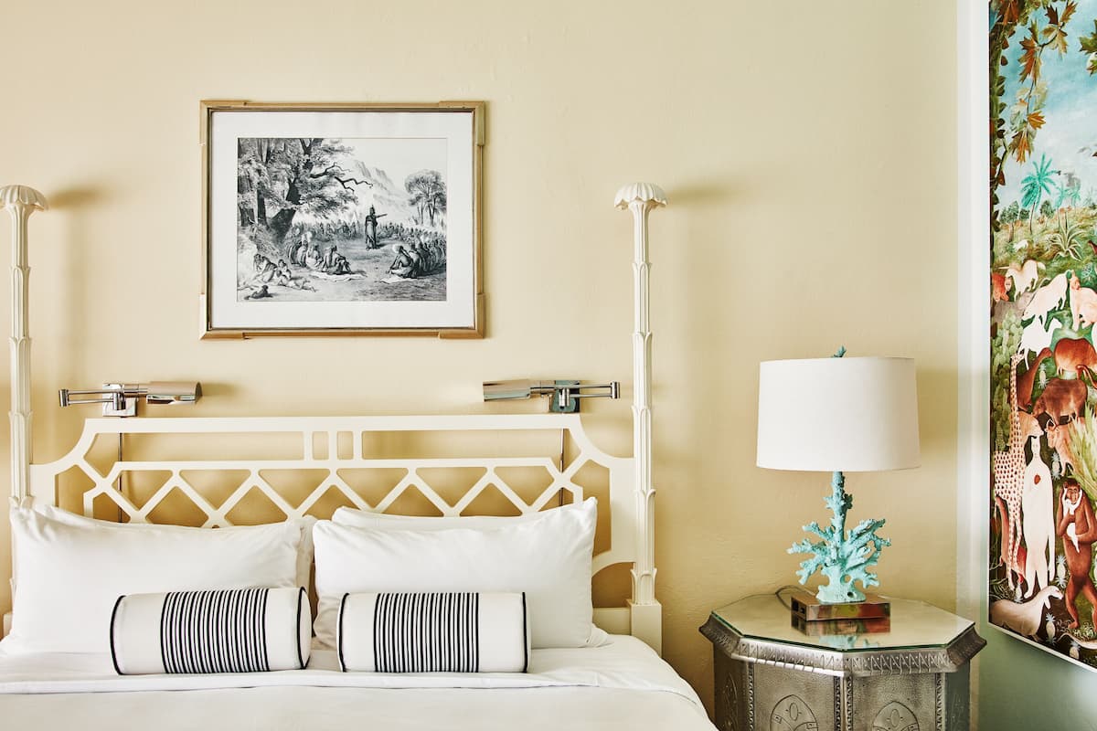 A hotel room with a bed, a framed black and white landscape painting, a white lamp, and a small table with a decorative item.