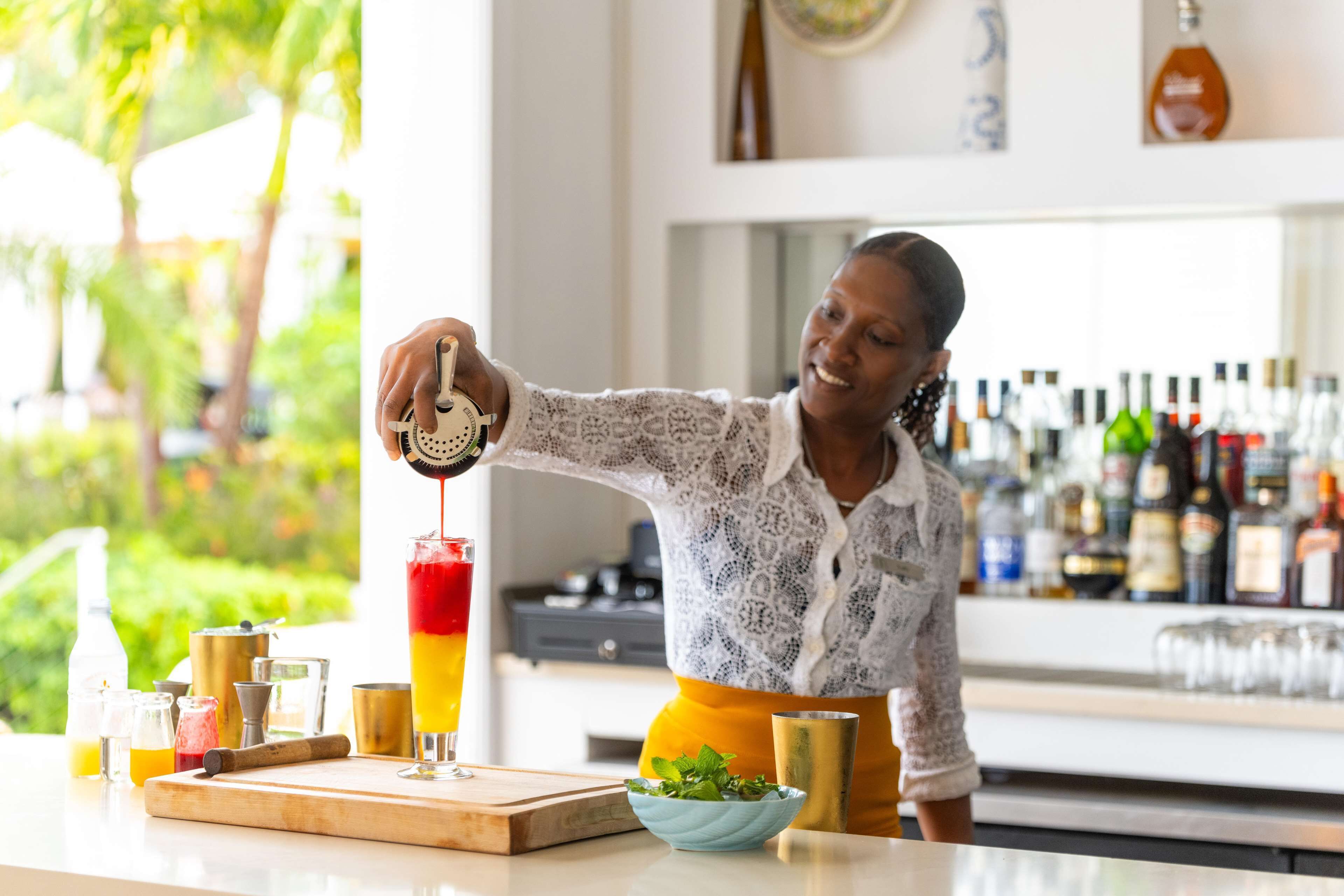 A bar setting with a bartender preparing a drink, a bar counter with various bottles, and a window with a view of greenery.
