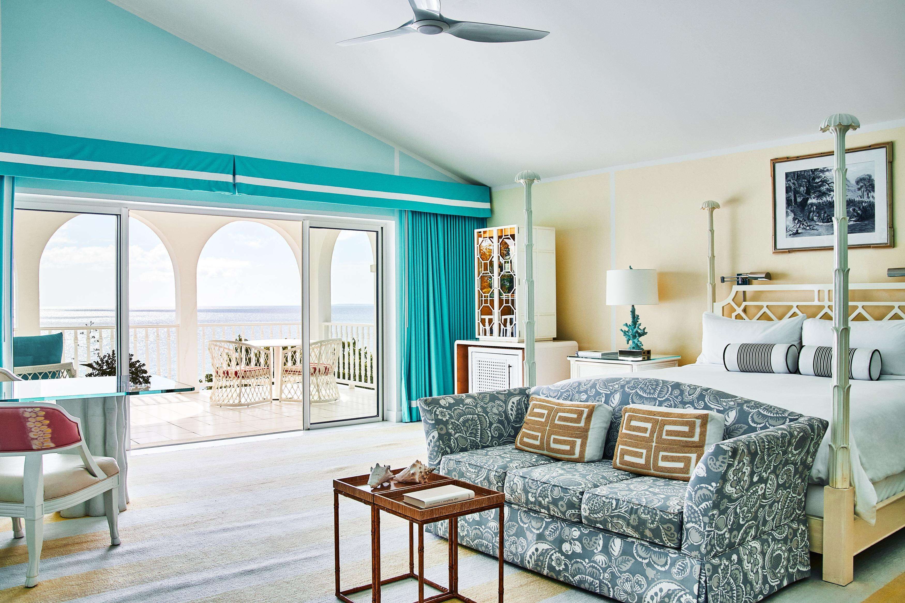 A hotel room with a balcony, a bed with patterned bedding, a sofa, a small table, and a ceiling fan.