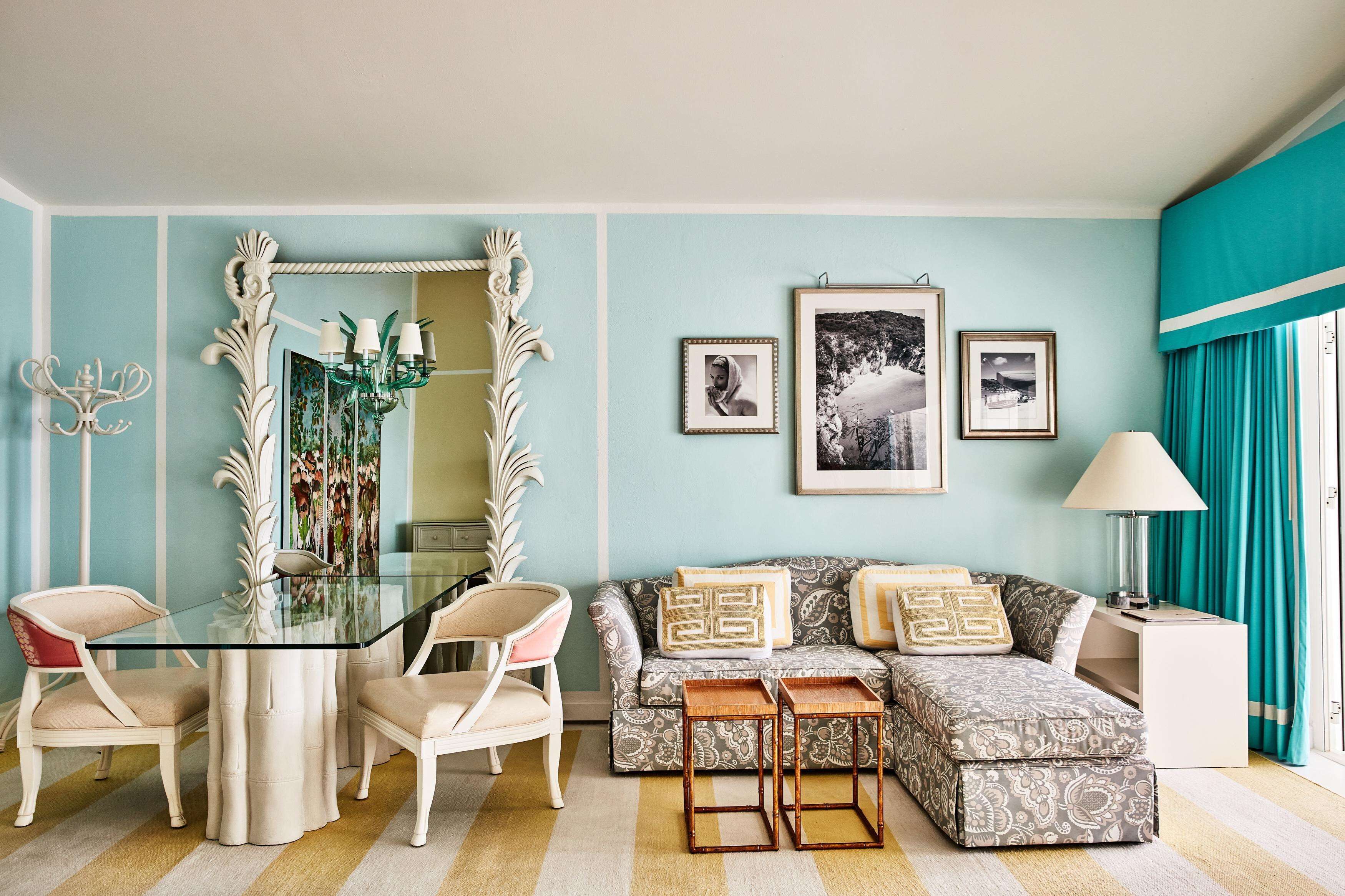 Une pièce comprenant une table à manger avec un plateau en verre, deux chaises, un canapé agrémenté de coussins à motifs, un miroir avec un cadre orné et une table d’appoint avec une lampe.