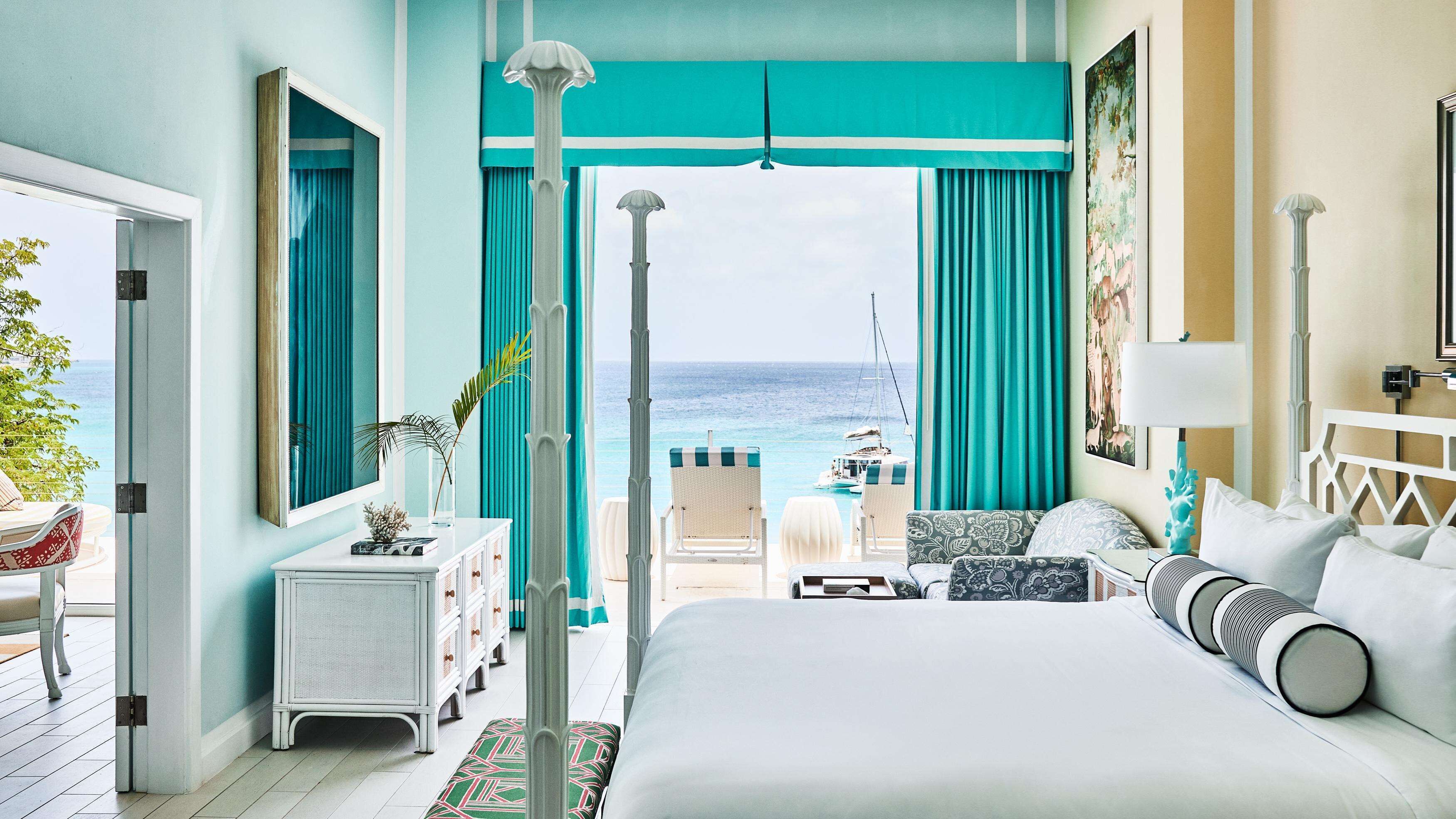 A bedroom with a large bed, a view of the ocean, a balcony with a table and chairs, and a door leading outside.