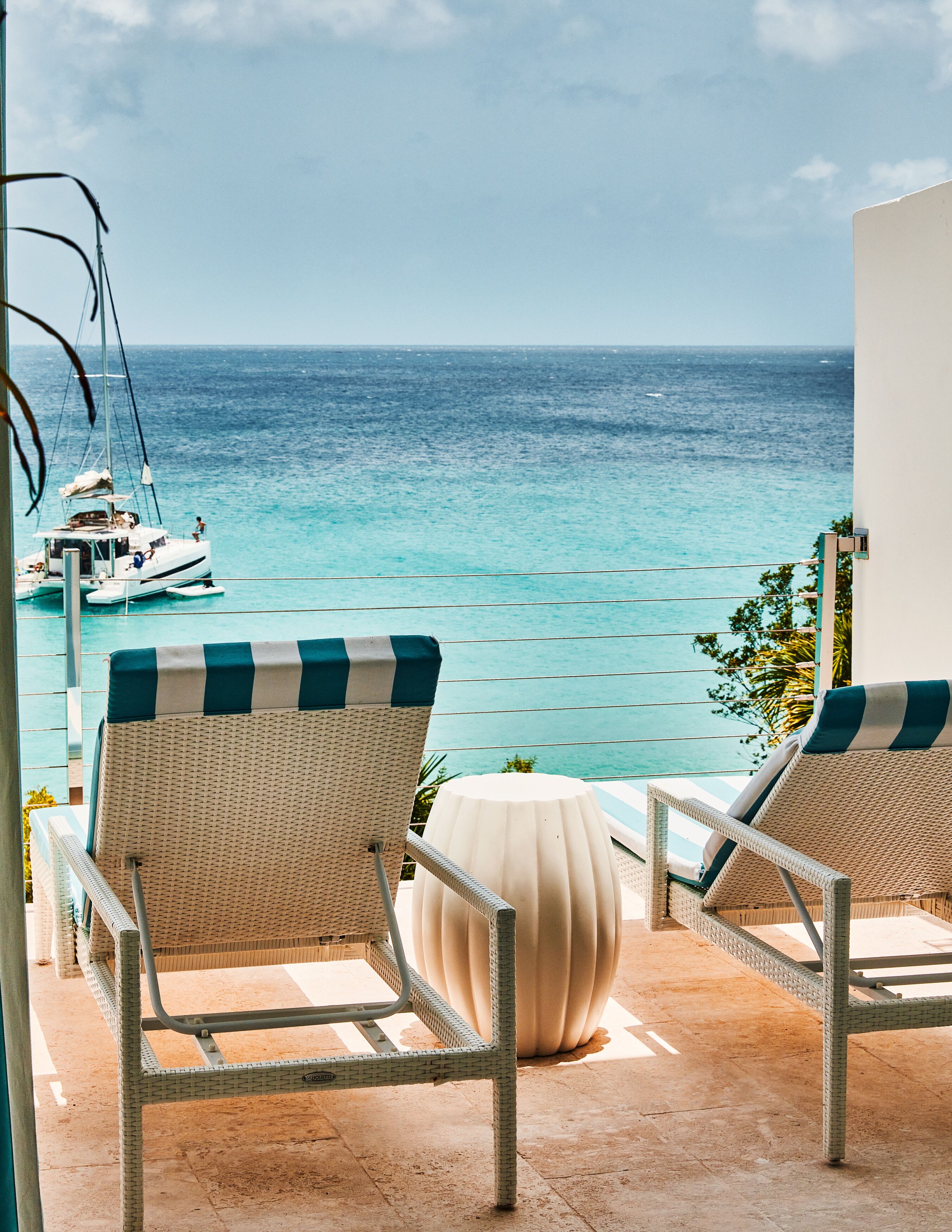 Two wicker lounge chairs with striped cushions, a round table, and a view of the ocean with a sailboat in the distance.