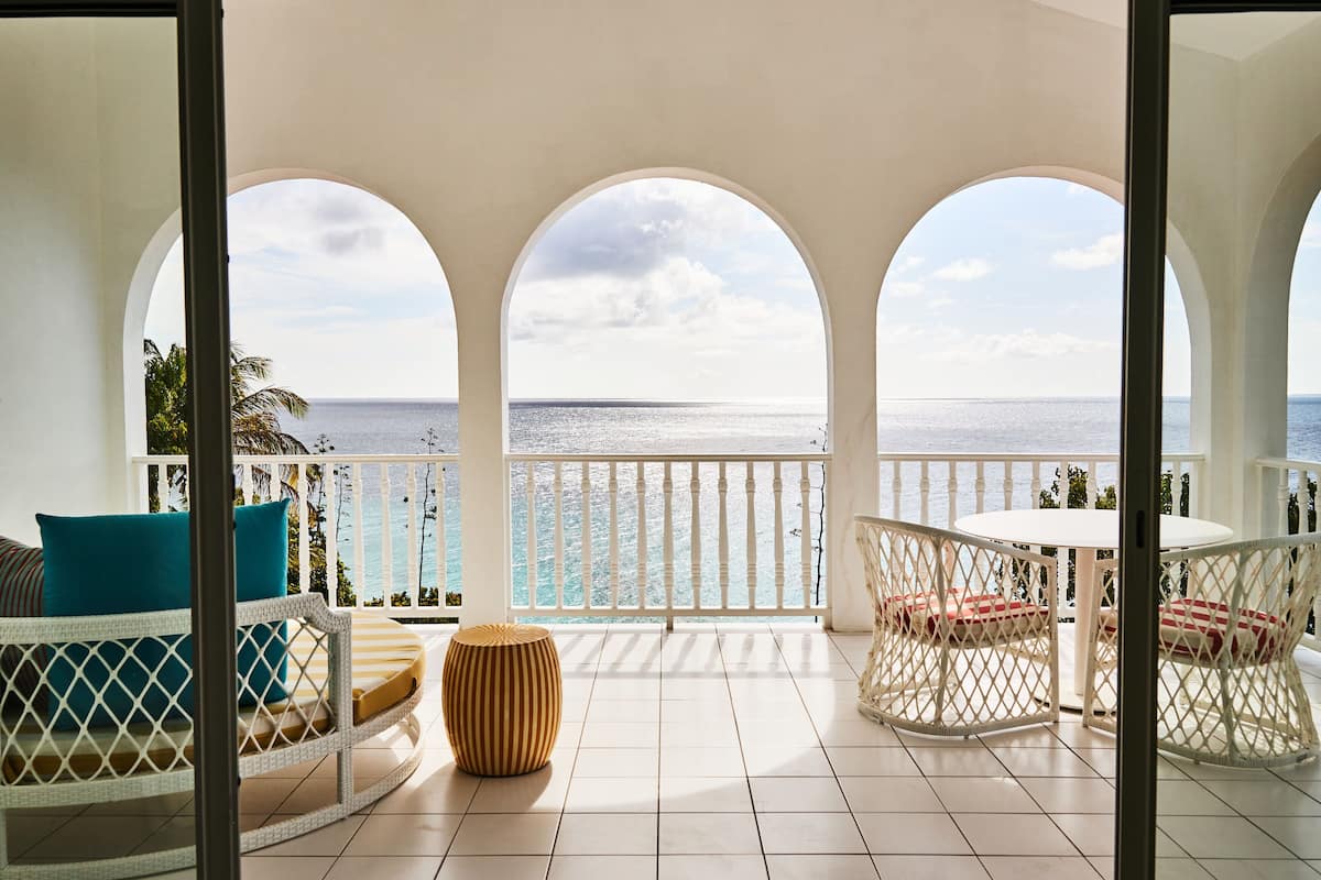 A balcony with a view of the ocean, furnished with a wicker sofa, a round table, and a small stool.