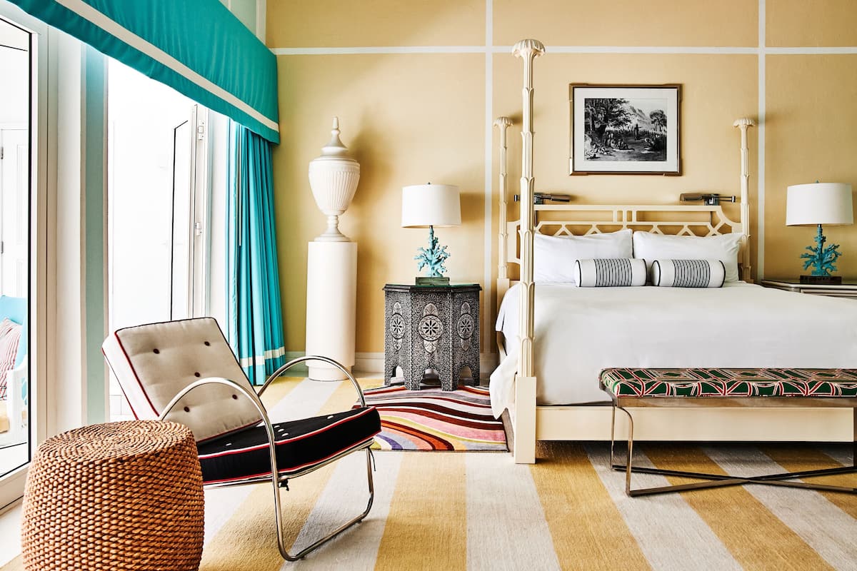 A hotel room with a four-poster bed, a chair, a small side table, and a large window with curtains.
