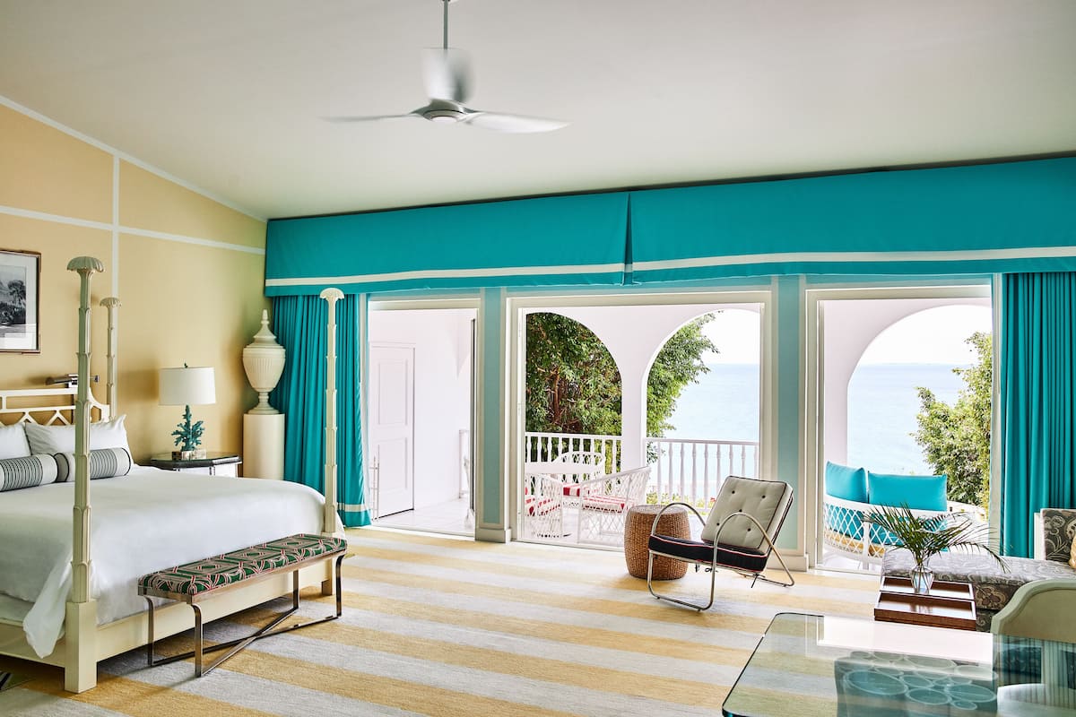 A spacious bedroom with a large bed, a sitting area with a glass table, and a balcony with a view of the sea.