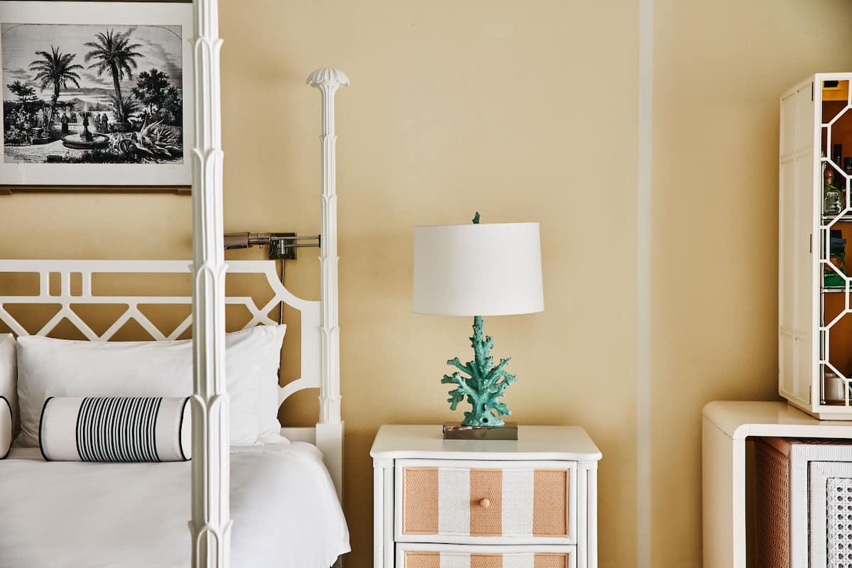 A bed with a white headboard, a nightstand with a white lamp, a framed picture on the wall, and a white cabinet with a geometric design.