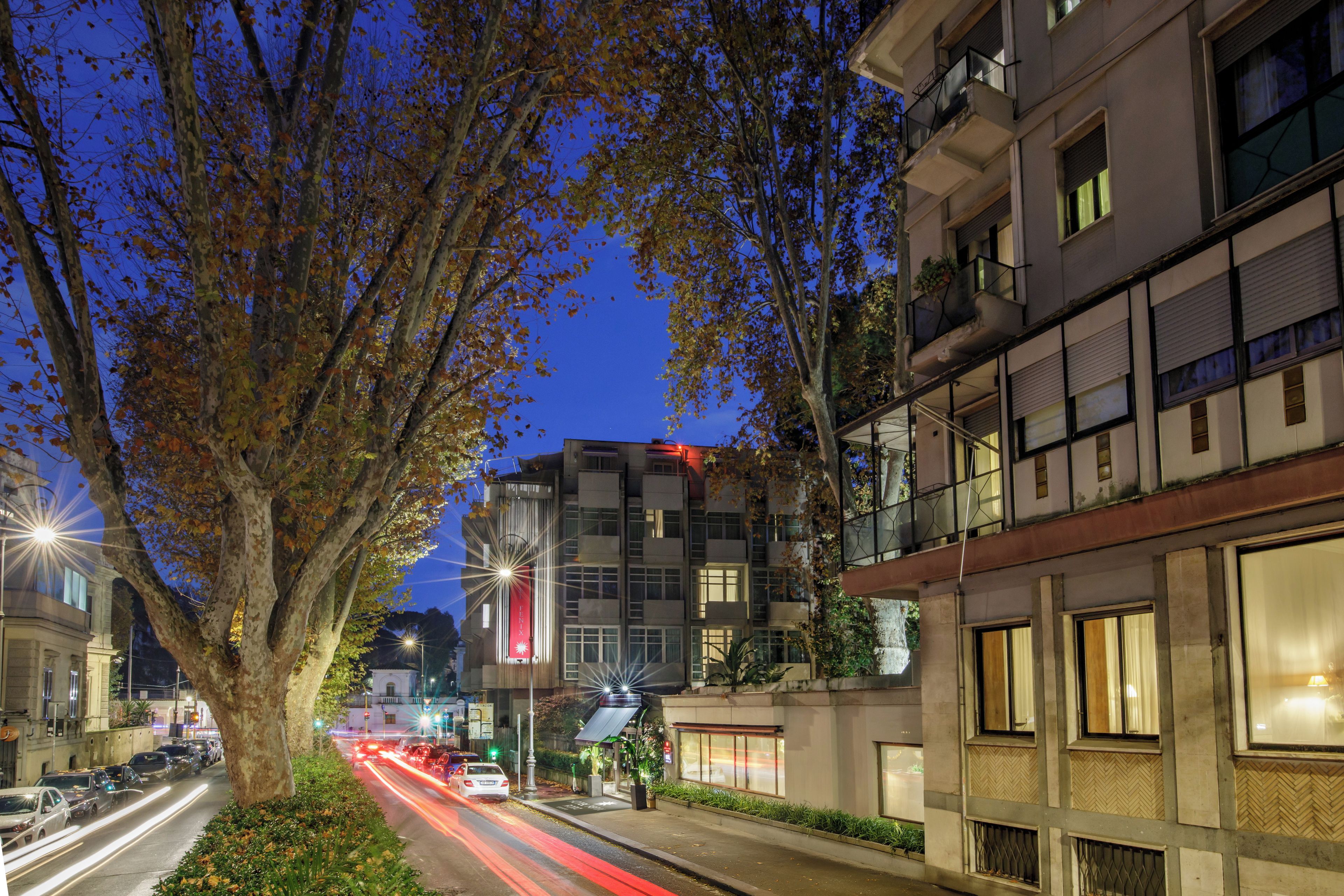 A tree-lined street with modern buildings, a visible sign for a hotel, and a clear view of the sky.