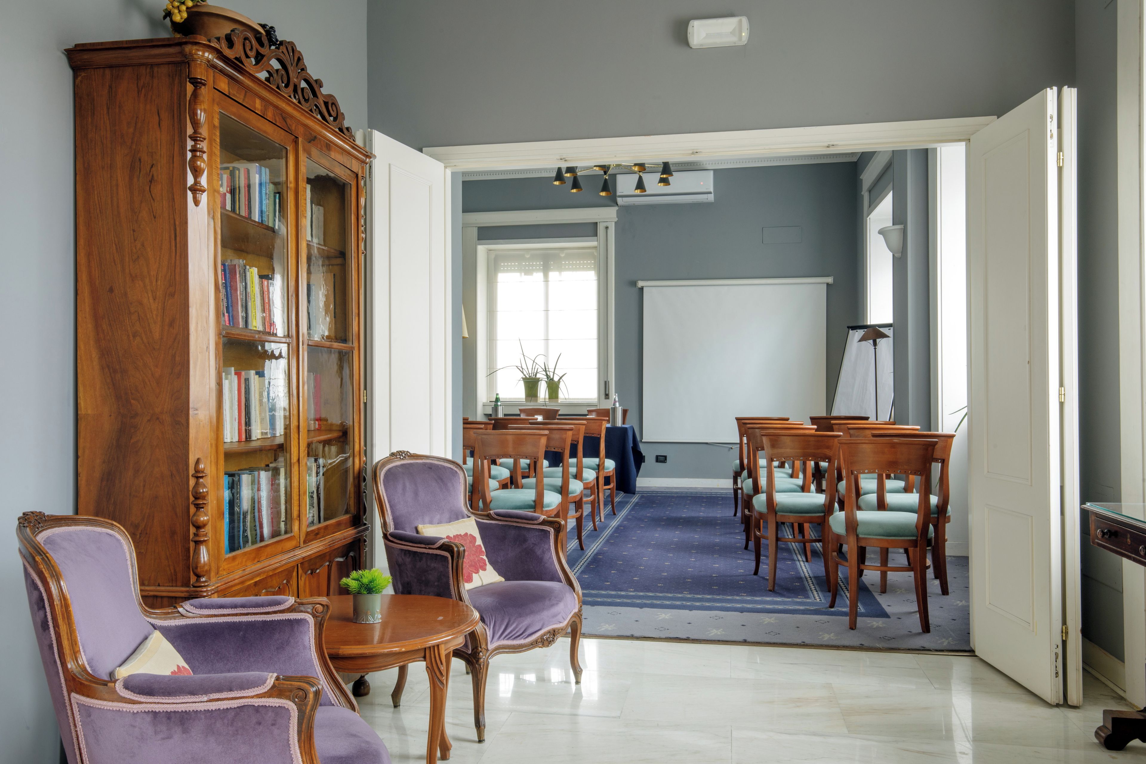 A room with a wooden bookcase, purple upholstered chairs, a round table, and a whiteboard.