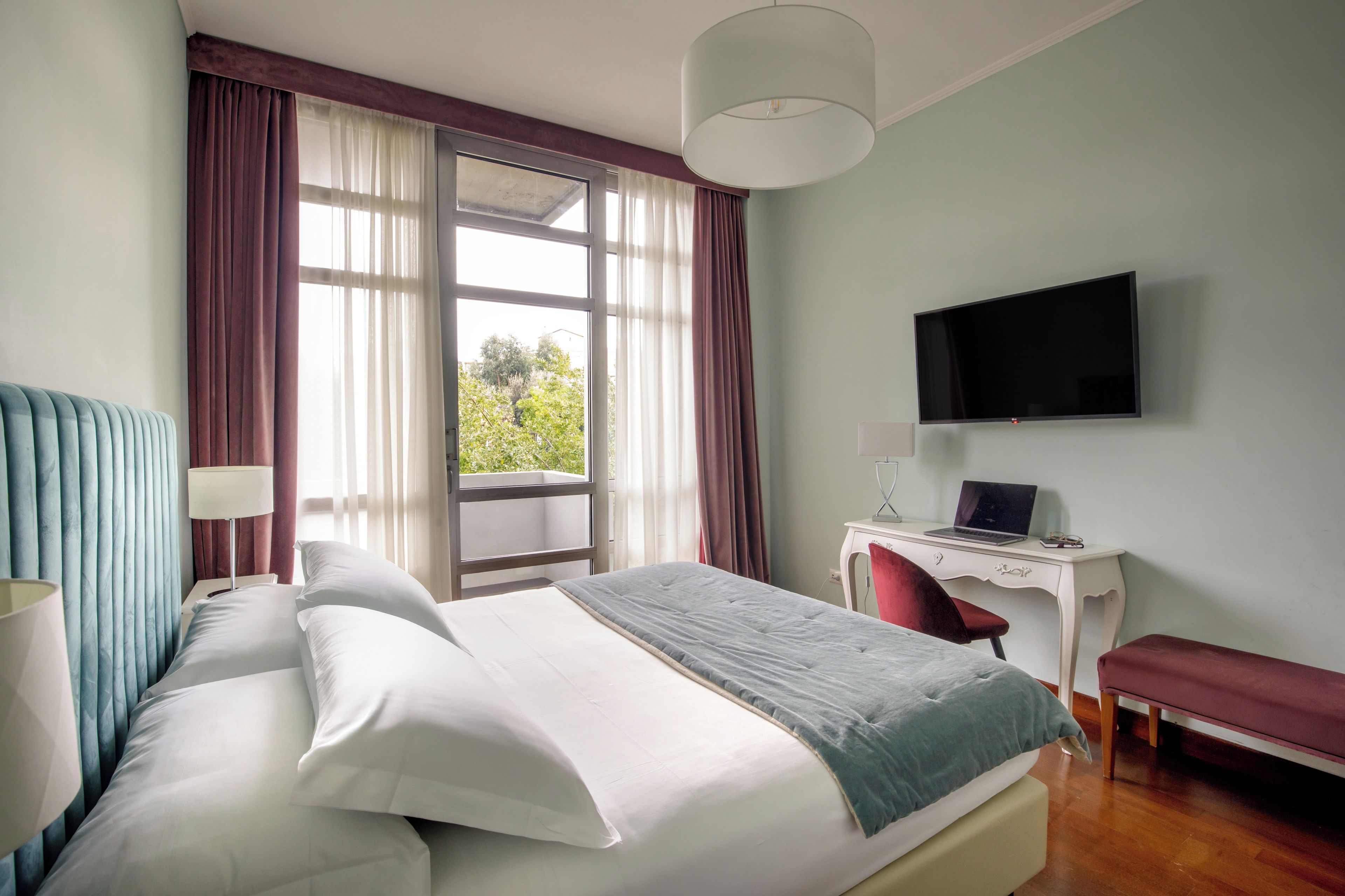 Ein Hotelzimmer mit einem Bett, einem Schreibtisch mit Laptop, einem Fernseher und einem Fenster mit Vorhängen.