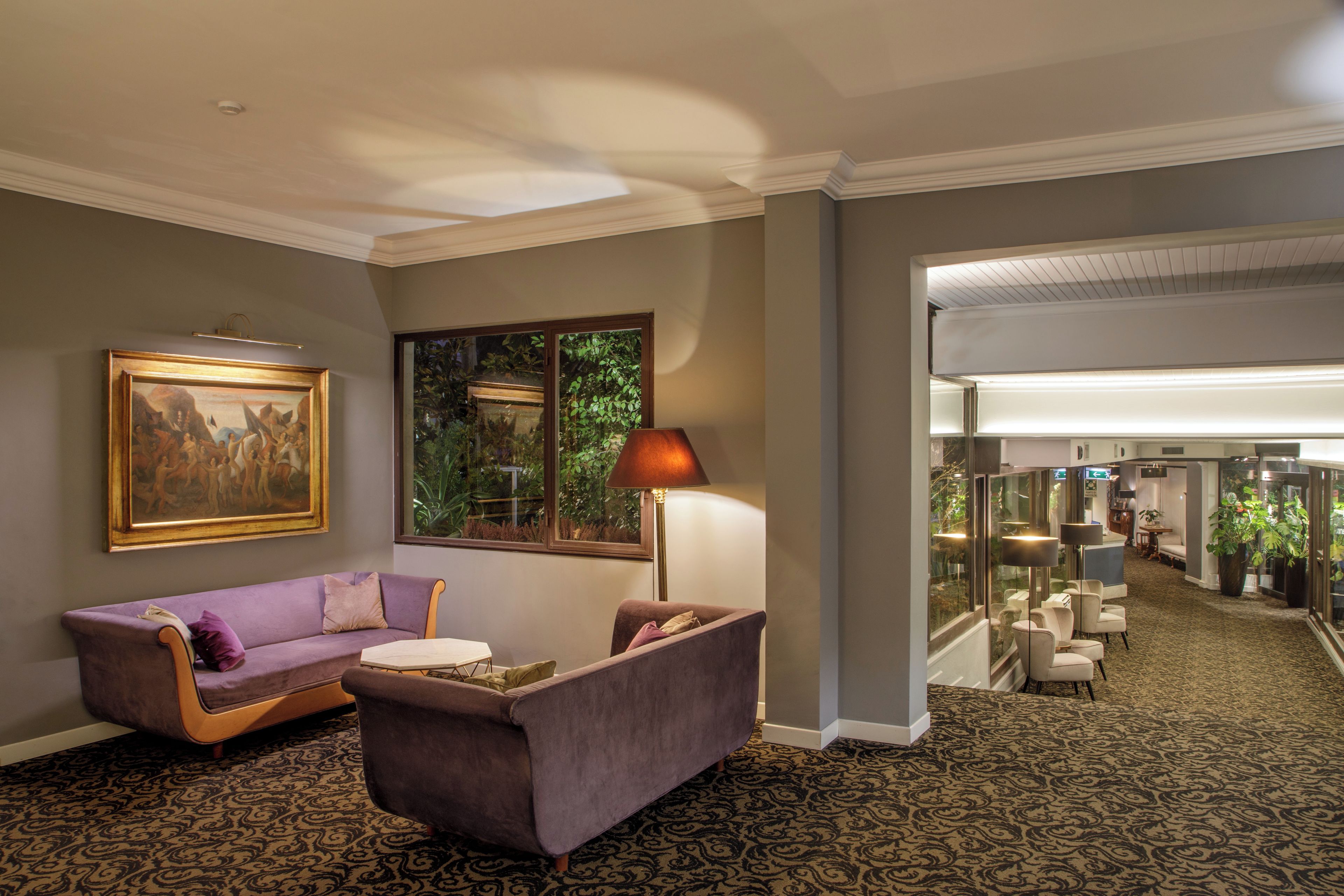 A lobby with a purple sofa, a table, a lamp, a painting, and a view of greenery through a window.