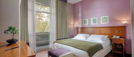 A hotel room with a bed, bedside table, lamp, and a view of trees through the window.