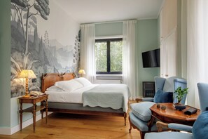 Un dormitorio con cama, mesita de noche con lámpara, una silla, televisor y ventana con cortinas.