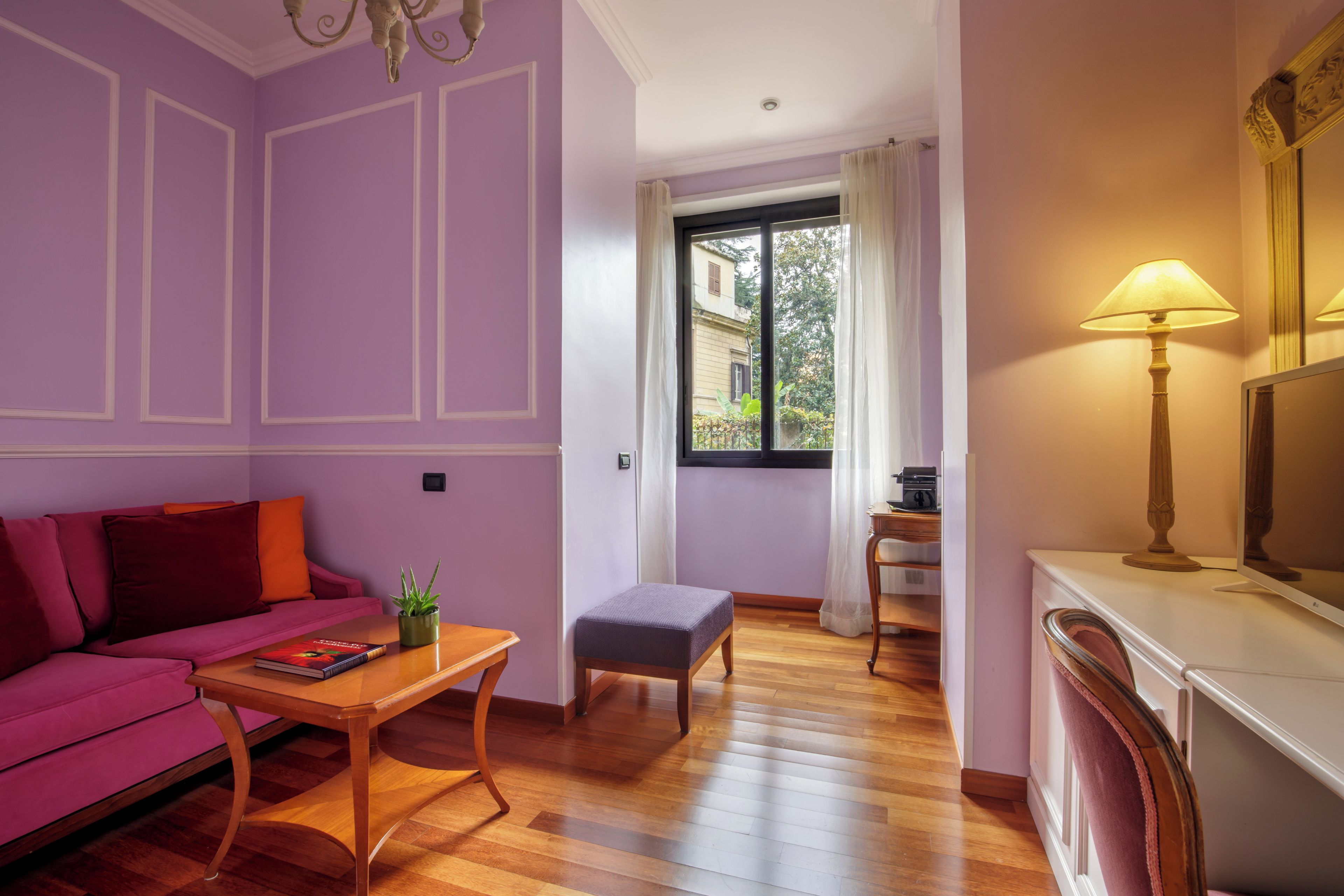 A room with a purple wall, a wooden floor, a pink sofa, a small table with a book, a window with a view of buildings, and a lamp.