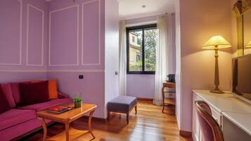 A room with a purple wall, a wooden floor, a pink sofa, a small table with a book, a window with a view of buildings, and a lamp.