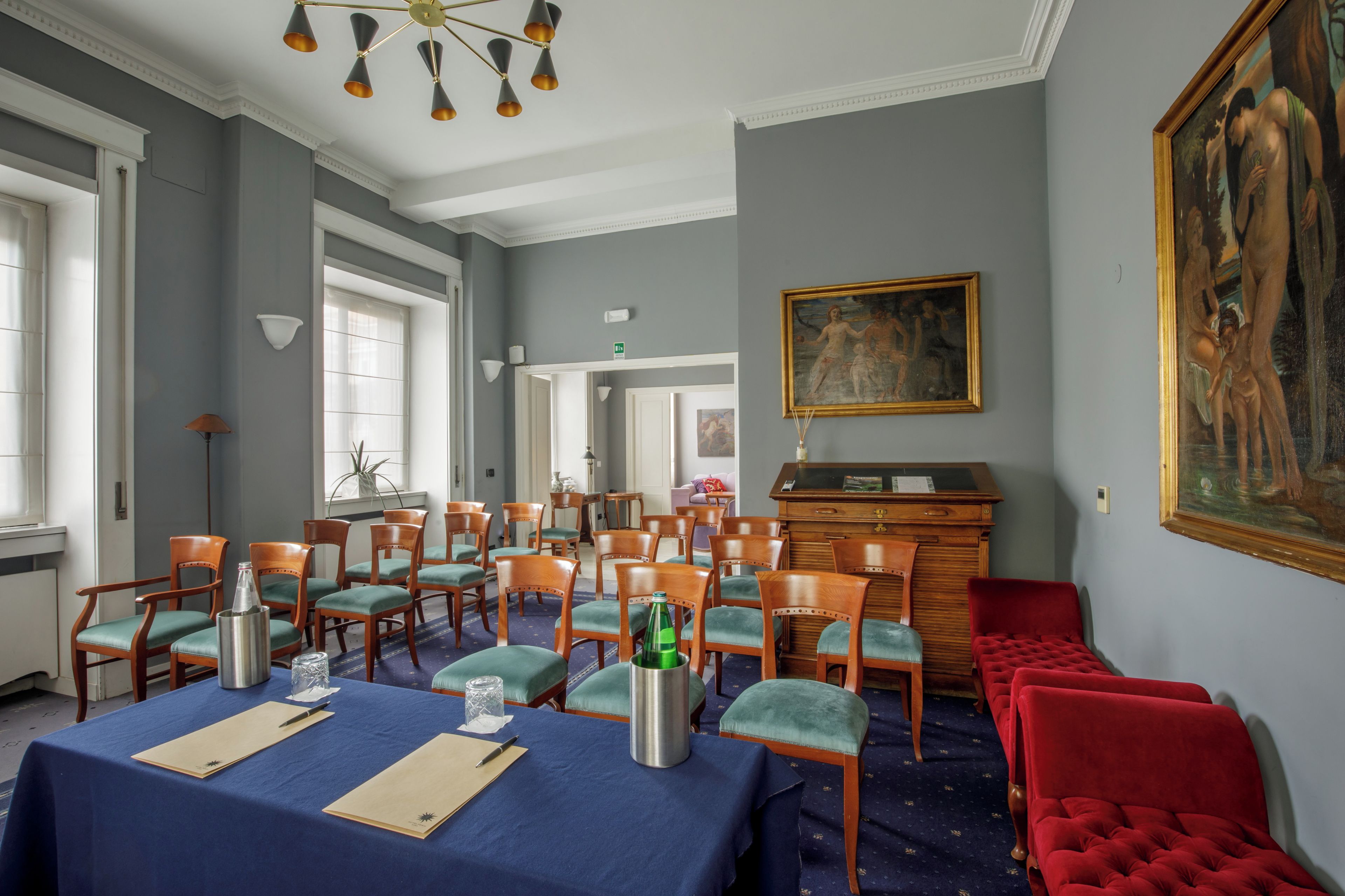 Quarto com uma mesa azul posta para refeições, cadeiras de madeira, um sofá vermelho e um quadro na parede.