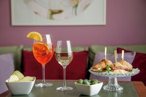 A dining table with a glass of red drink, a glass of clear drink, a plate of sandwiches, and bowls of chips and olives. - Fenix Hotel (Rome)