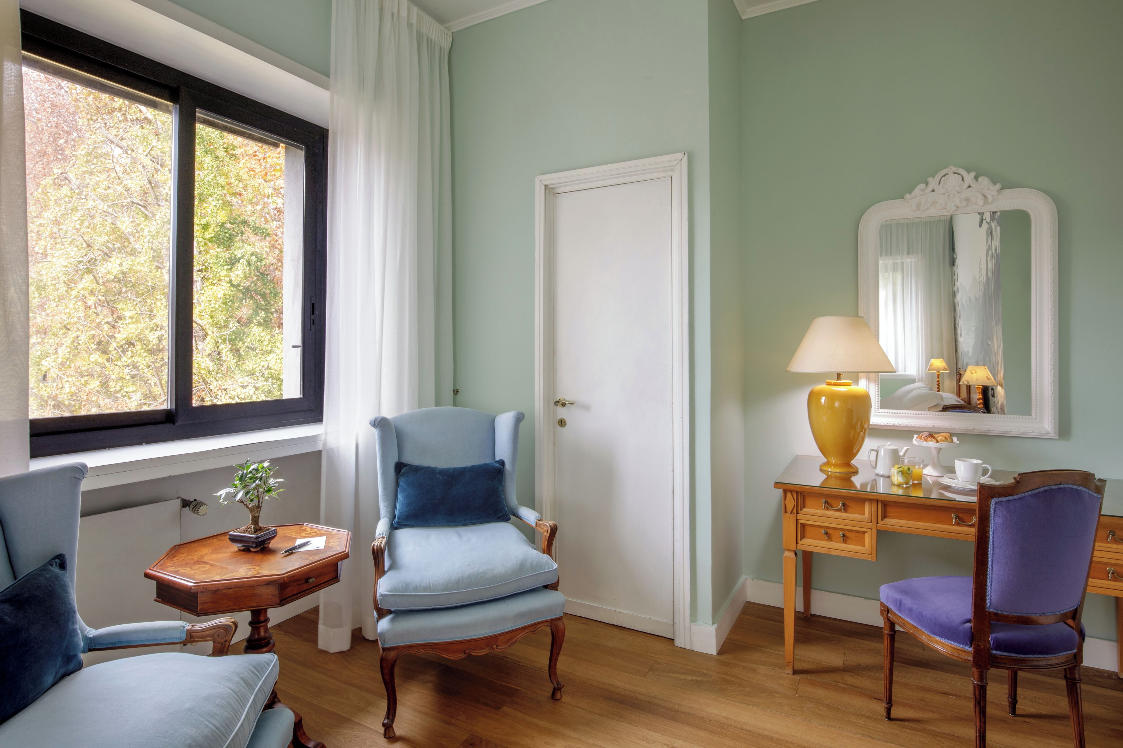 Ein Zimmer mit Holzboden, einem blauen Sessel, einem kleinen Tisch mit Pflanze, einem Schreibtisch mit Lampe und einem Spiegel.