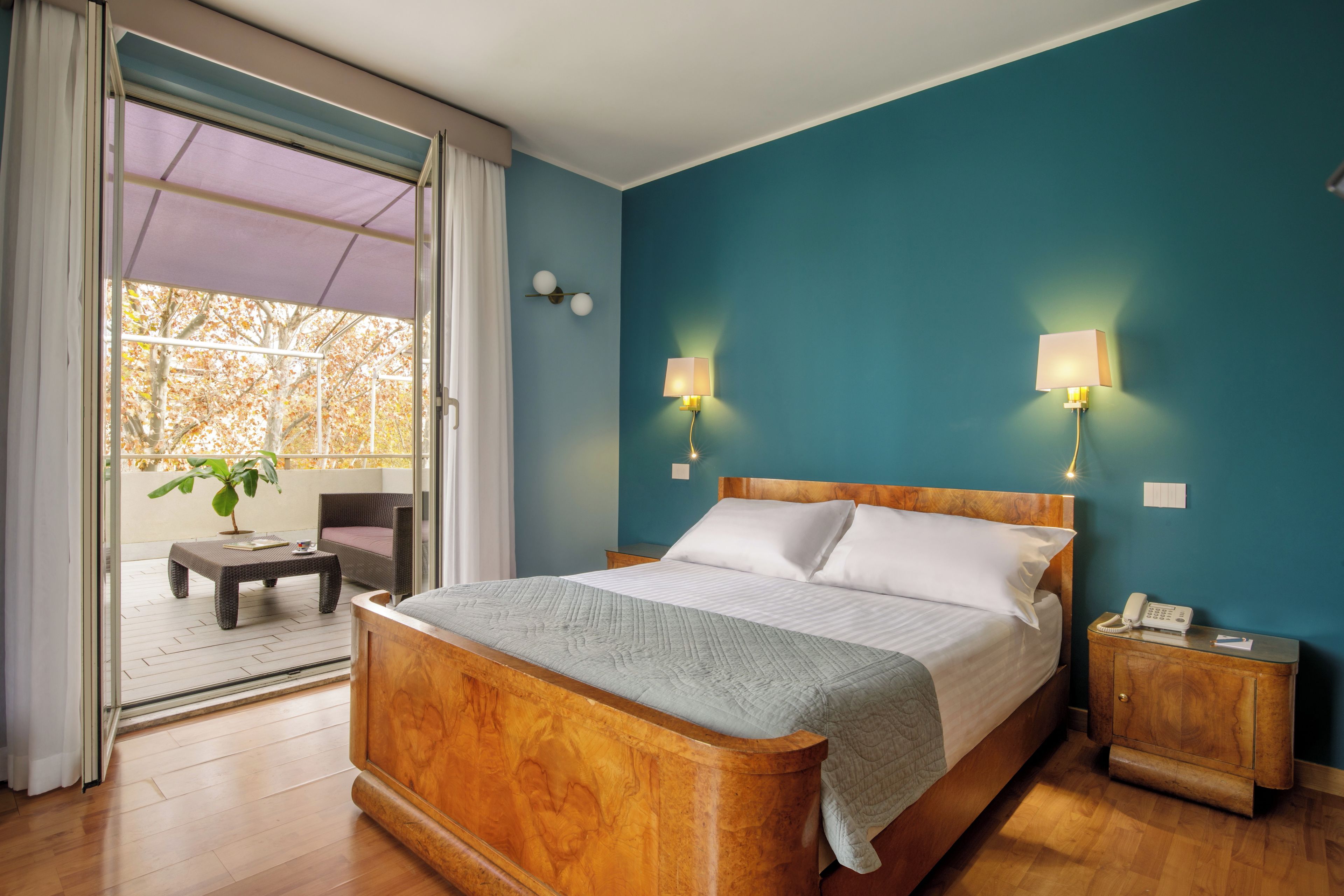A hotel room with a wooden bed, a small bedside table, a nightstand with a phone, and a view of the outdoors through a sliding glass door.