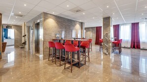 Un hall d’hôtel moderne avec un sol en marbre, un bar central doté de chaises rouges et un escalier menant à un autre niveau.
