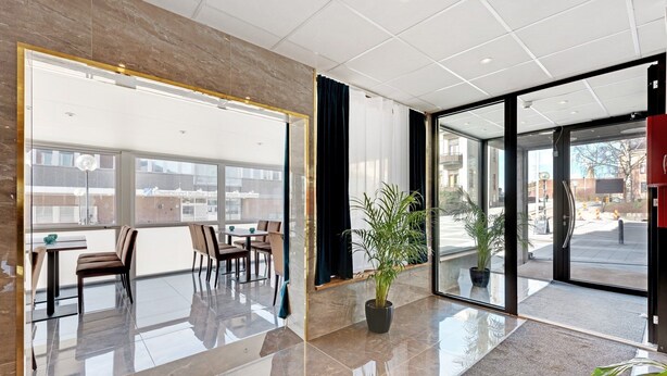Un hall d’hôtel moderne avec une cloison en verre, un espace salon avec des tables et des chaises, et une plante en pot.