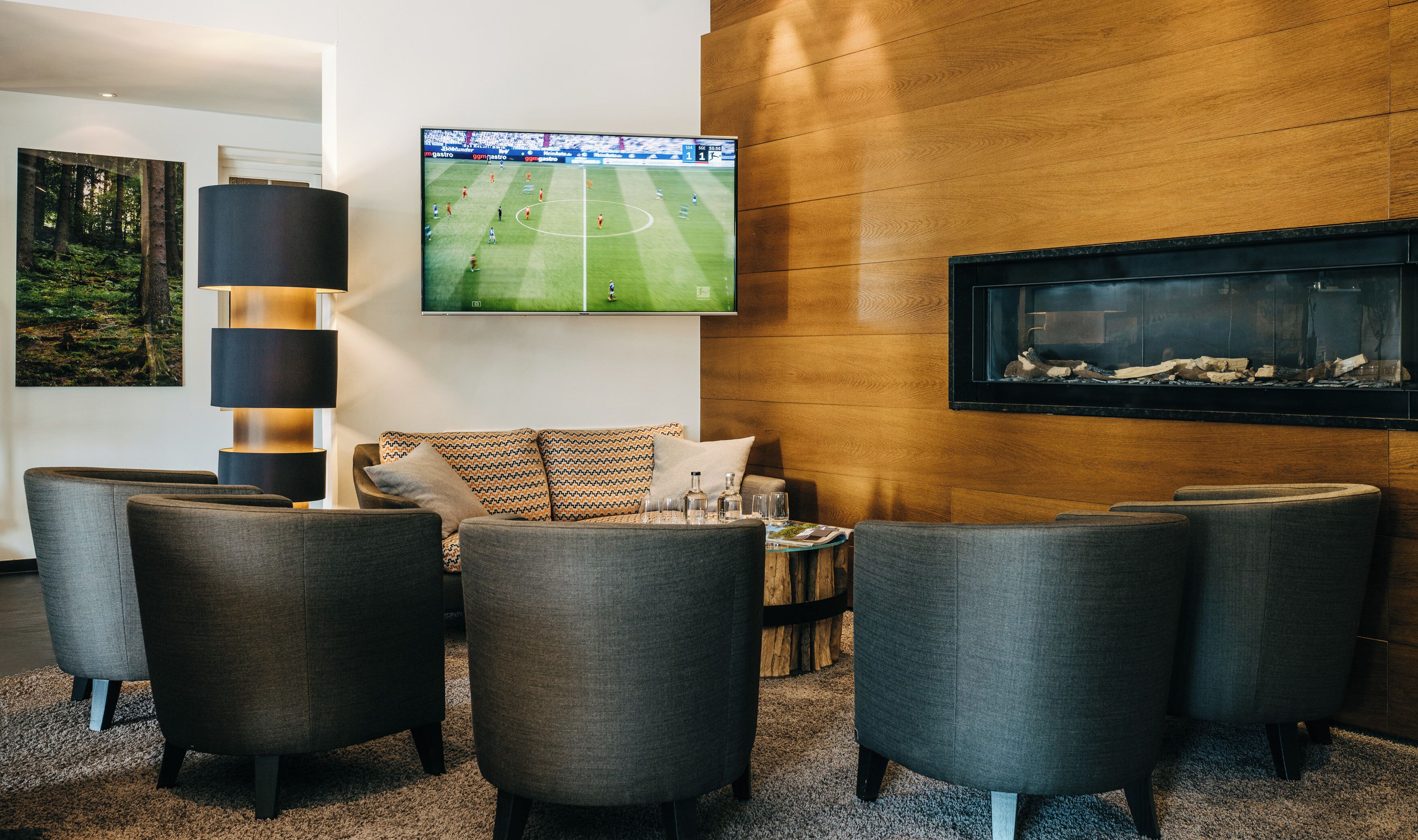 A modern lounge area with a large flat-screen TV displaying a soccer match, comfortable seating, a fireplace, and a framed picture on the wall.