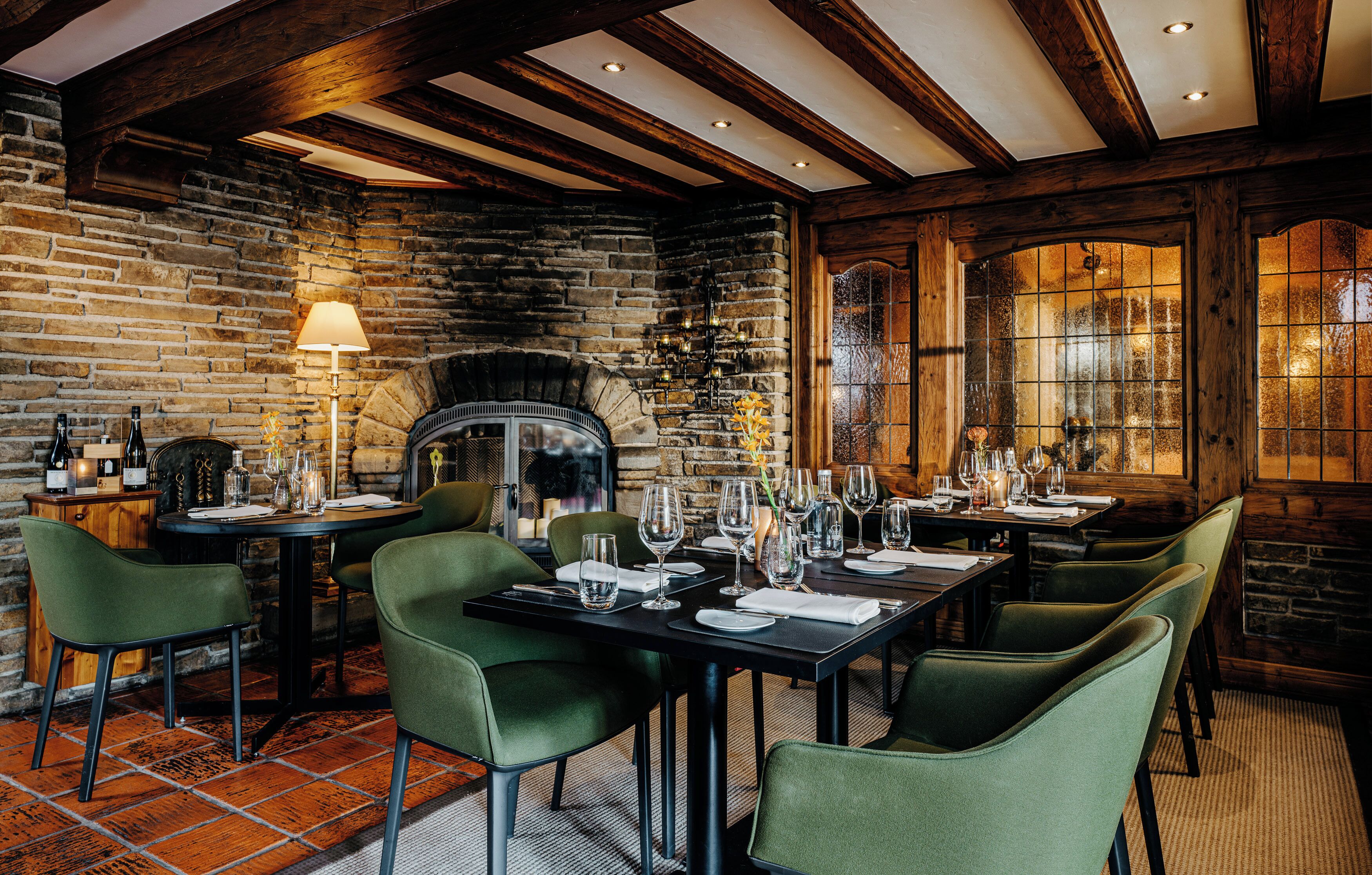 A restaurant with brick walls, wooden beams, and green upholstered chairs. Tables are set with wine glasses and plates.