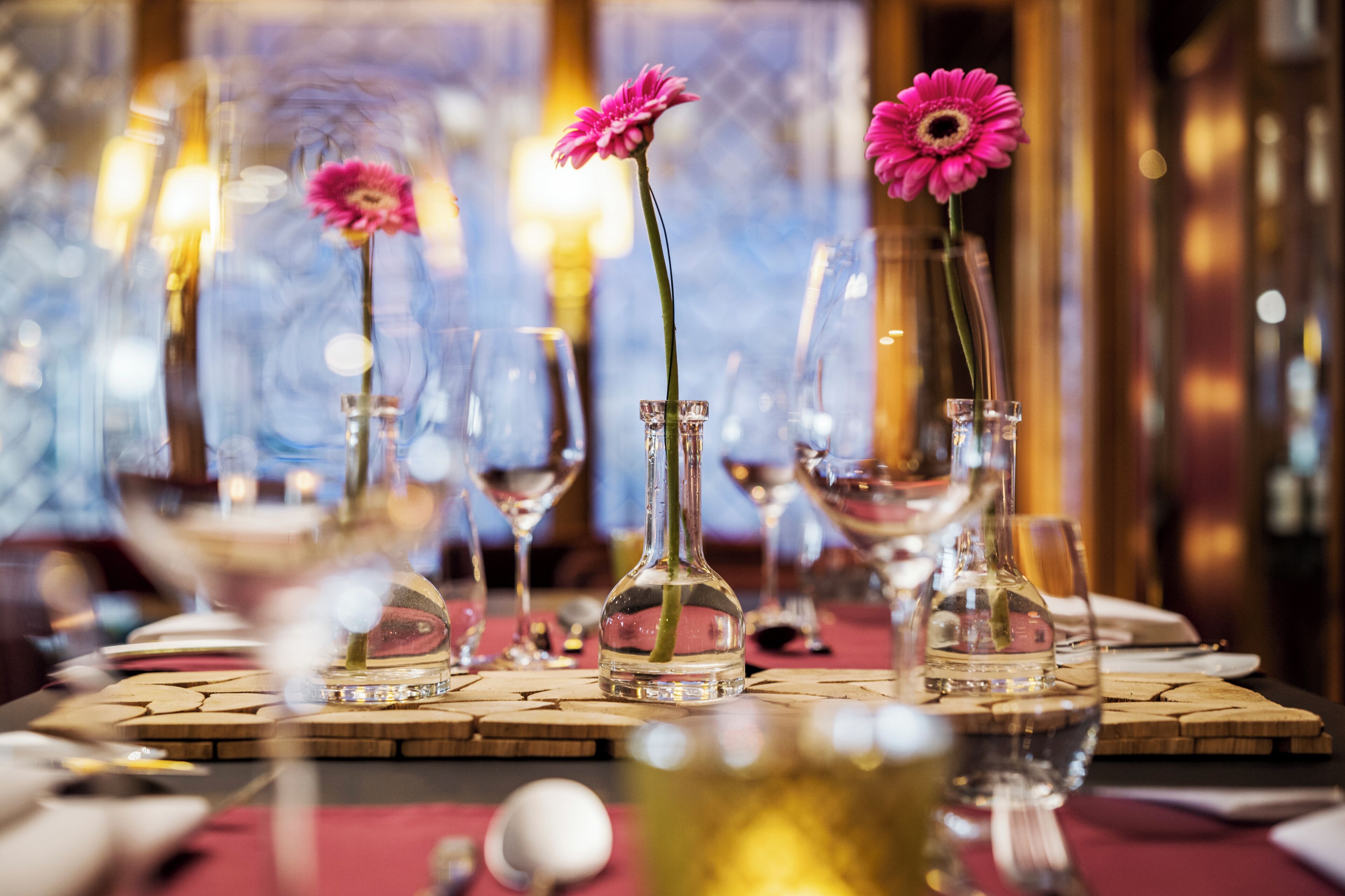 A dining table with clear glass vases containing pink flowers, surrounded by wine glasses and a wooden table runner.