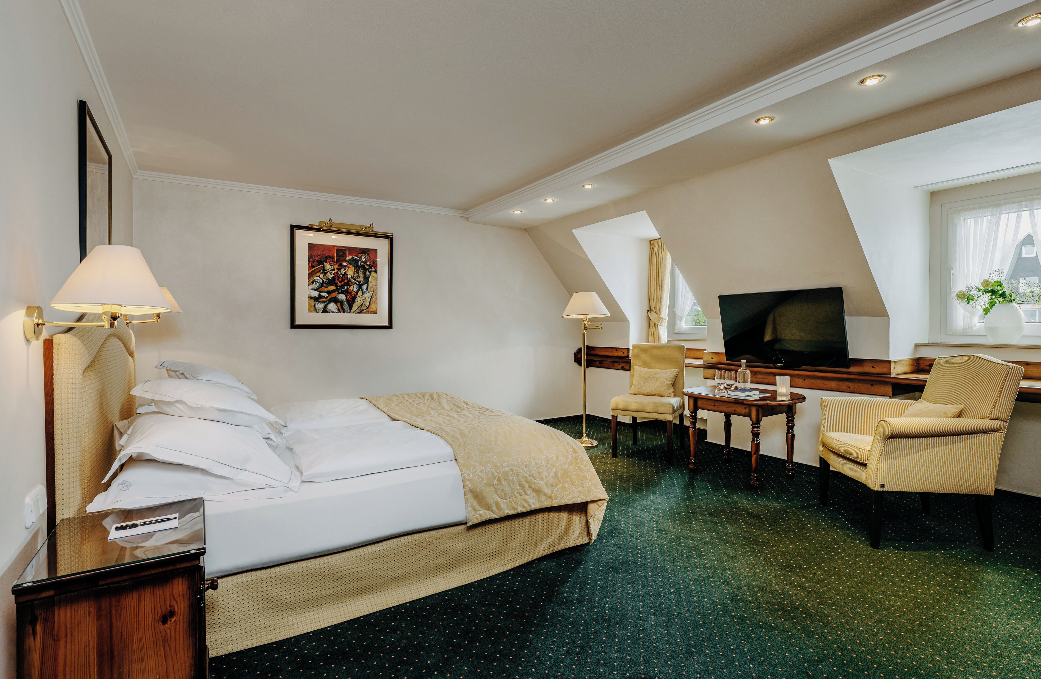 Habitación de hotel con cama, dos sillas, una mesita, televisor y un cuadro en la pared.