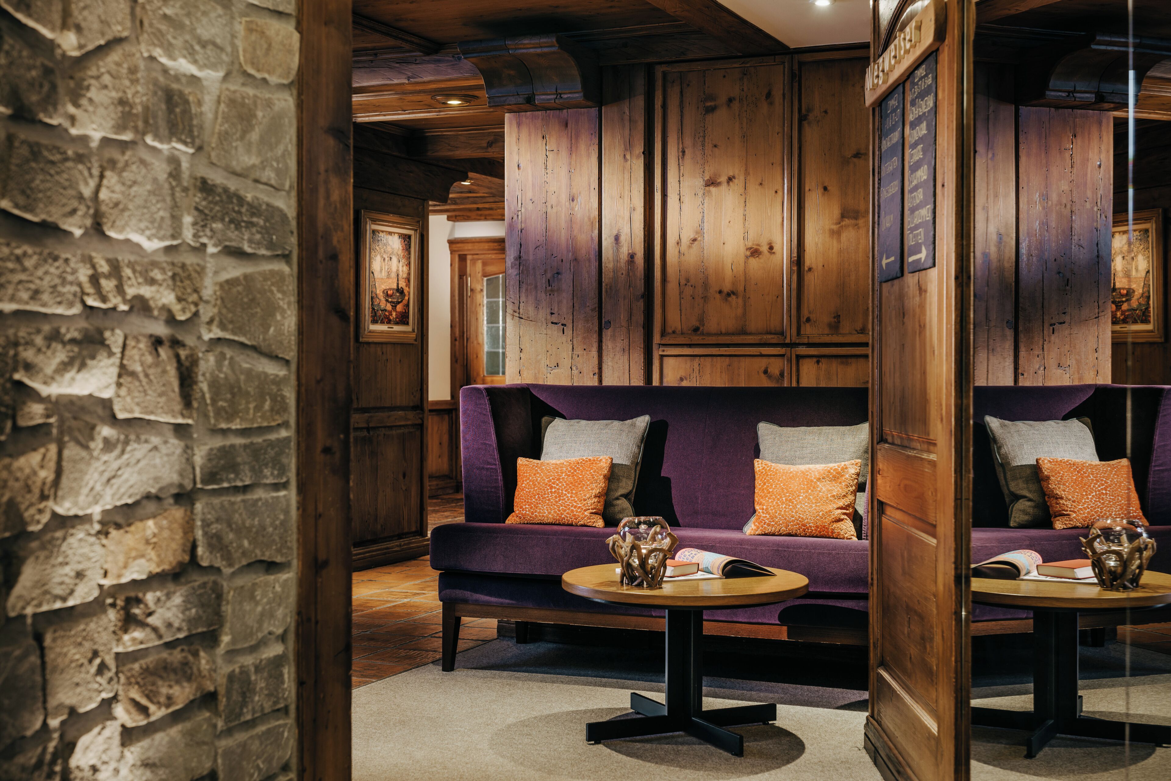 Ein Zimmer mit Steinwand, Holzvertäfelung, einem violetten Sofa mit orangefarbenen Kissen und zwei runden Tischen mit Dekoration.