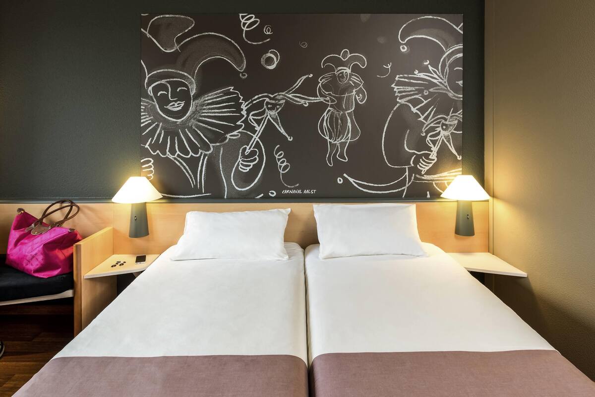 A hotel room with two beds, a nightstand with a lamp, a wall with a chalkboard drawing, and a pink bag on the left side.