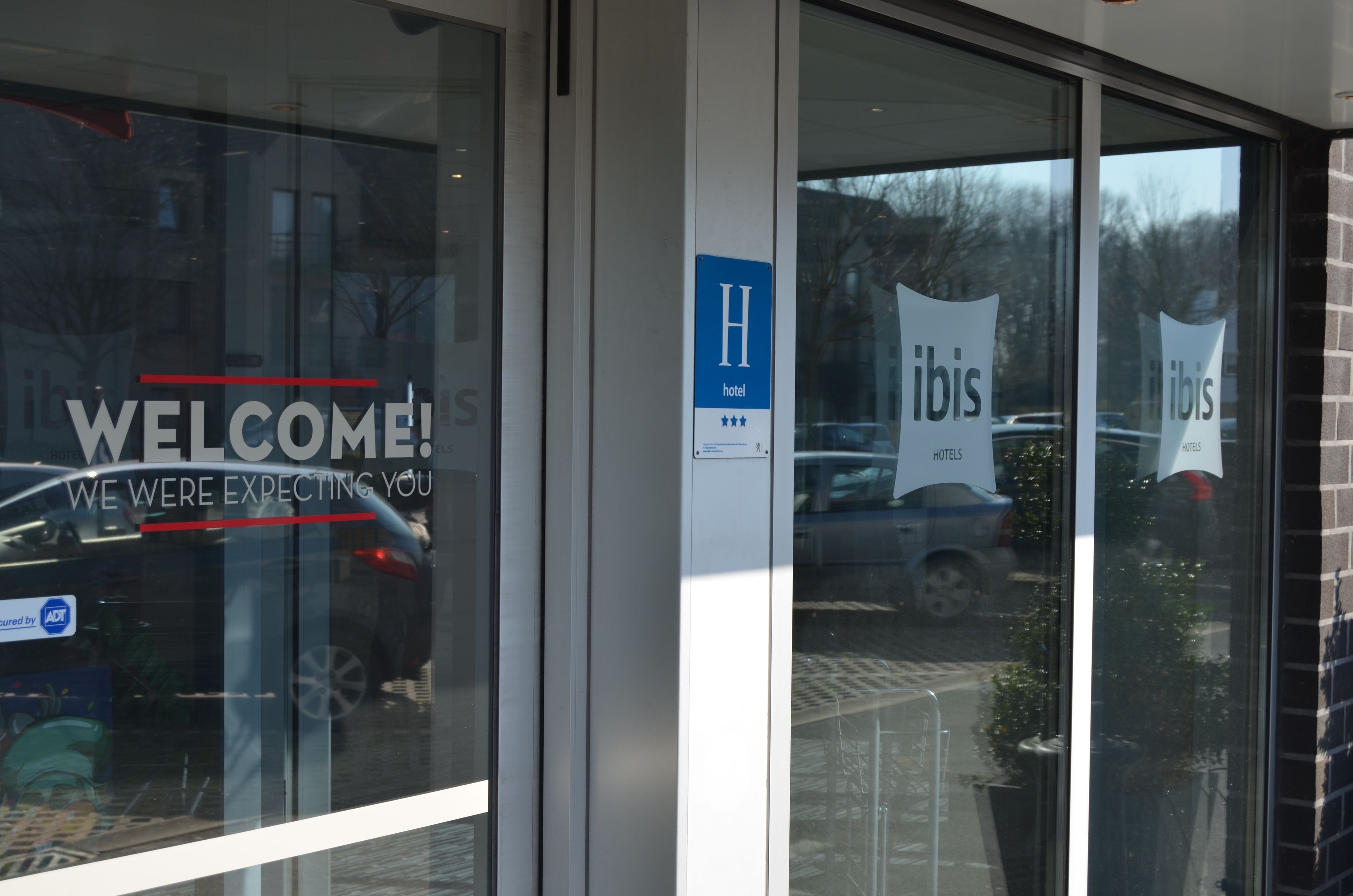 A hotel entrance with a 'WELCOME!' sign, a 'H' sign for the hotel, and 'ibis hotels' banners.