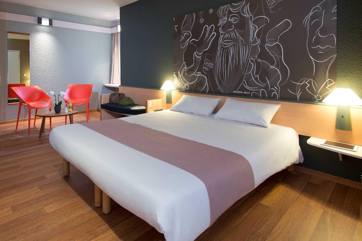 A hotel room with a large bed, two bedside lamps, a nightstand, a chair, a small table with a plant, and a wall with a mural.