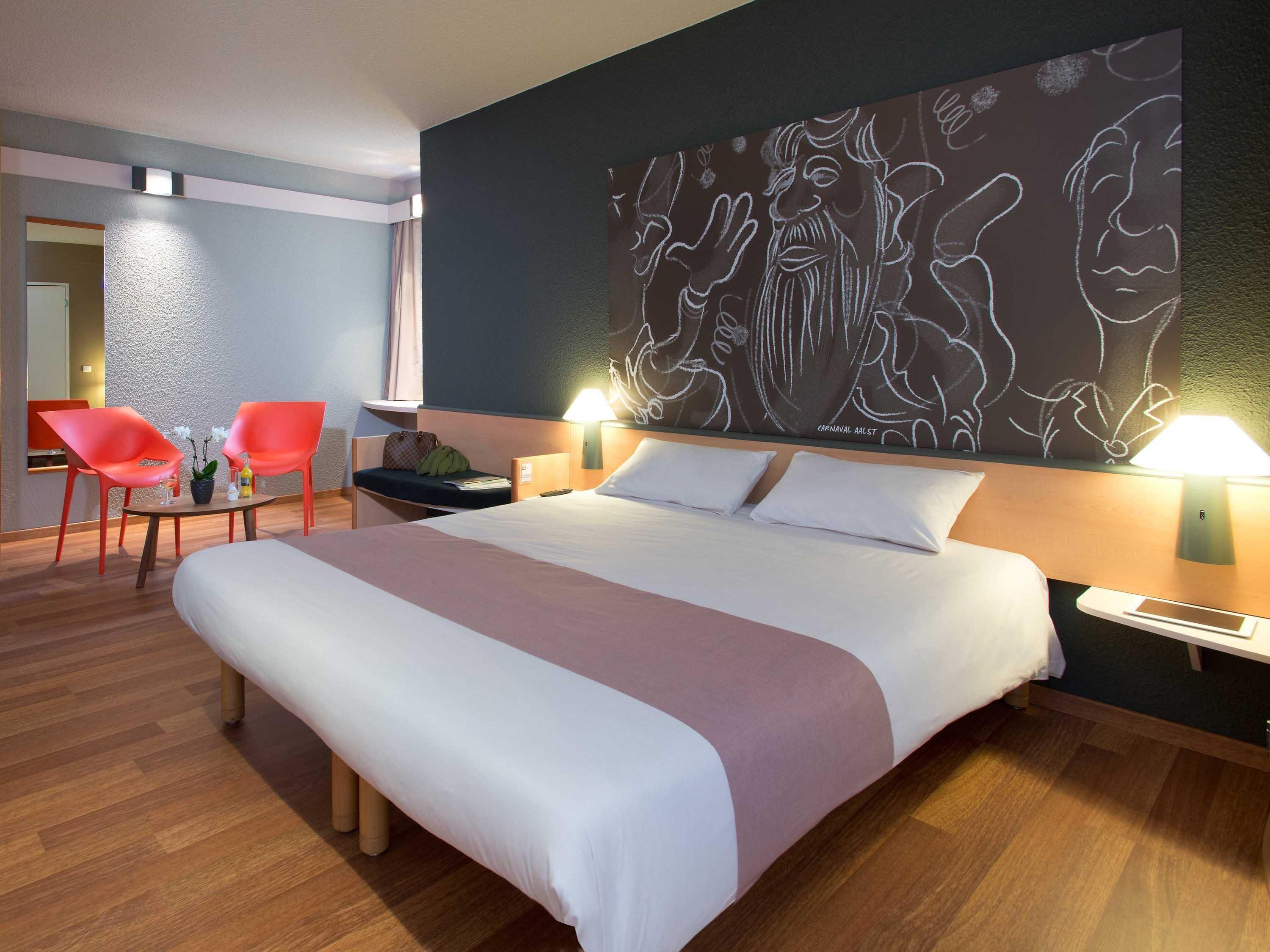 Une chambre d’hôtel avec un grand lit, deux lampes de chevet, une table de chevet, une chaise, une petite table avec une plante et un mur orné d’une fresque.