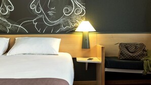 A hotel room with a bed, a nightstand with a lamp, a chair, and a wall art piece. - ibis Aalst Centrum (Aalst)