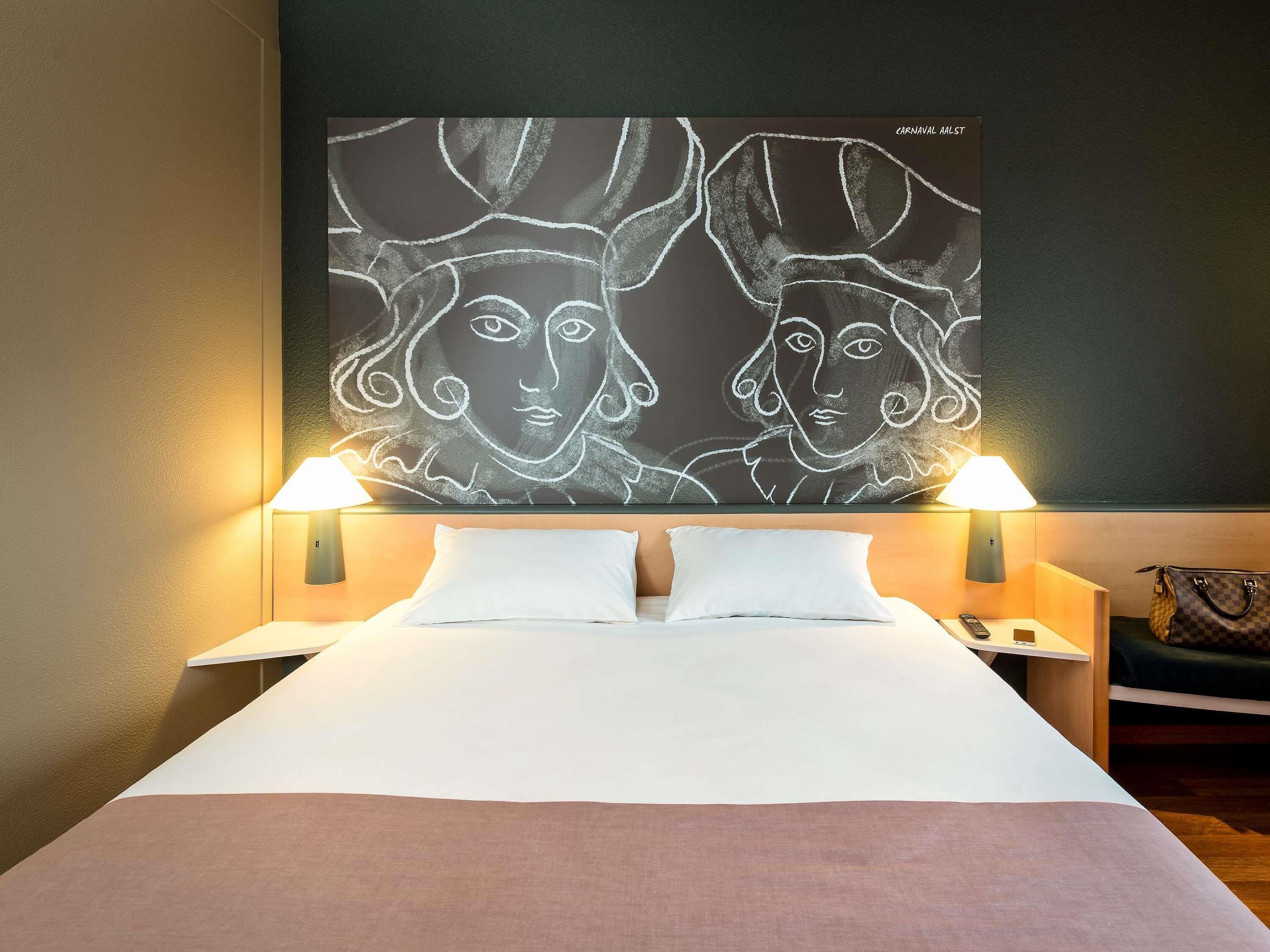 Habitación de hotel con cama, dos lámparas de noche, una mesita de noche, un bolso de mano y un cuadro abstracto de gran tamaño en la pared.