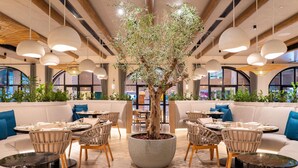 Un restaurante con mesas redondas, sillas de mimbre, un macetero central con un árbol y lámparas colgantes.