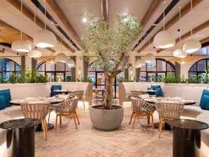 A restaurant with round tables, wicker chairs, a central tree planter, and hanging pendant lights. - ibis World Trade Centre Dubai (Dubai)