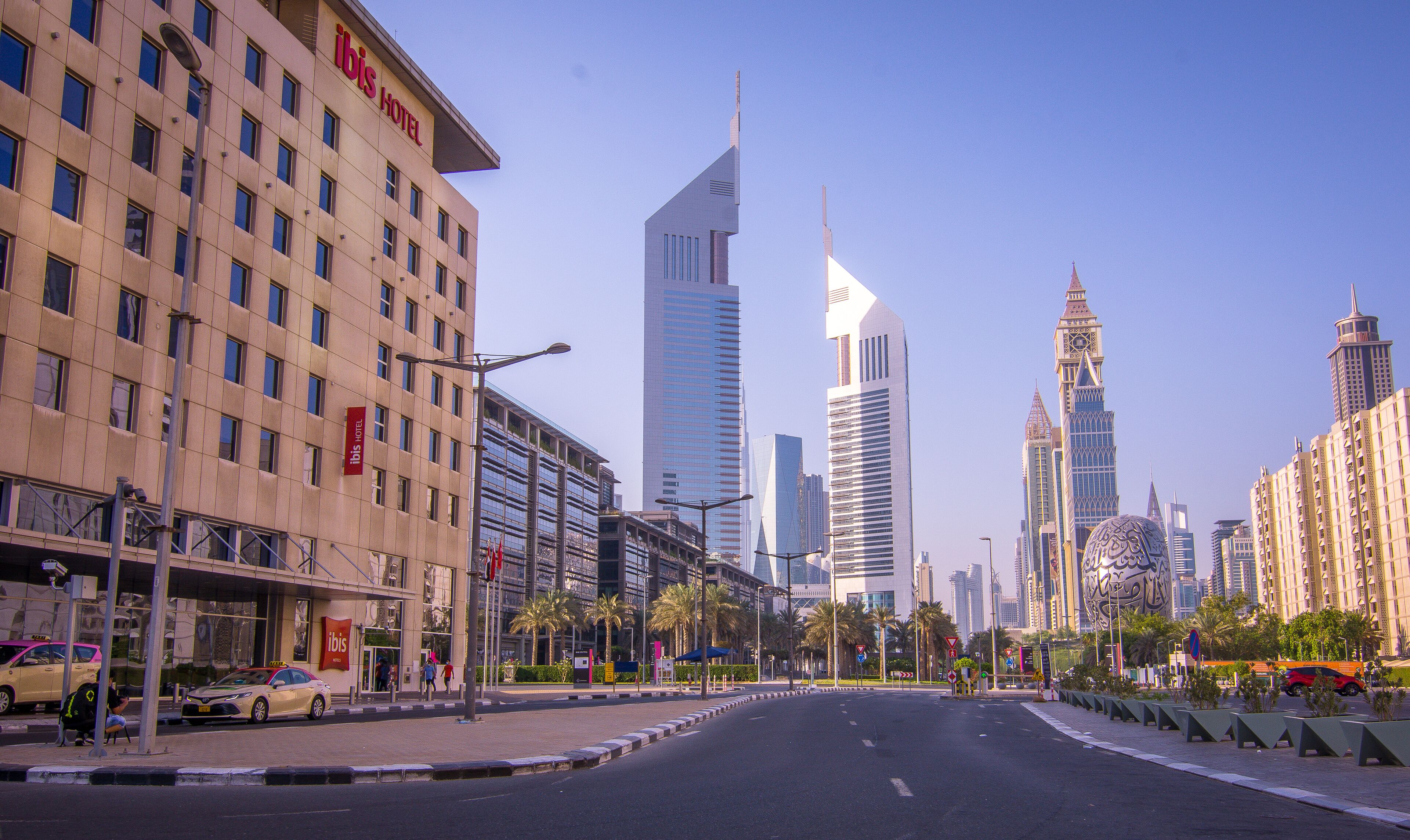 A modern hotel with a prominent "ibis Hotel" sign, surrounded by high-rise buildings and palm trees.