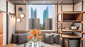Una habitación moderna con sofá, una mesita con jarrones, un mini refrigerador y una ventana con vista a la ciudad.