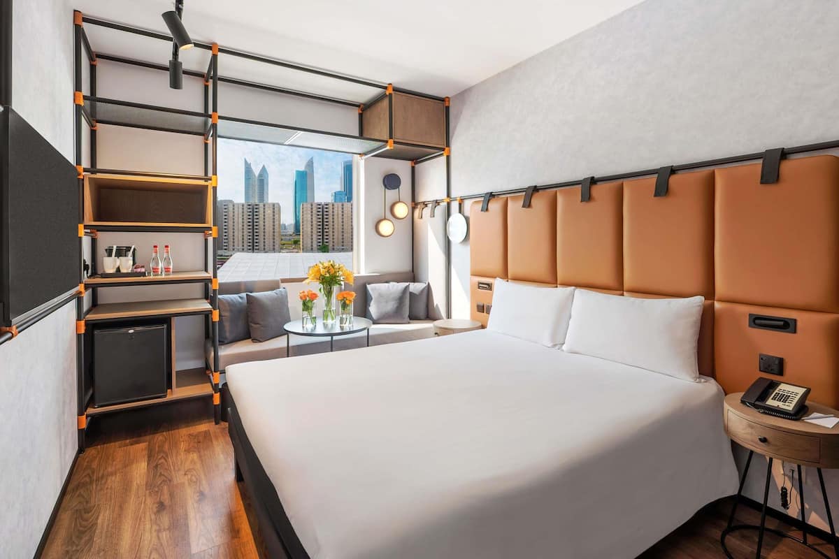 A modern hotel room with a large bed, a sofa, a small table with flowers, and a city view.