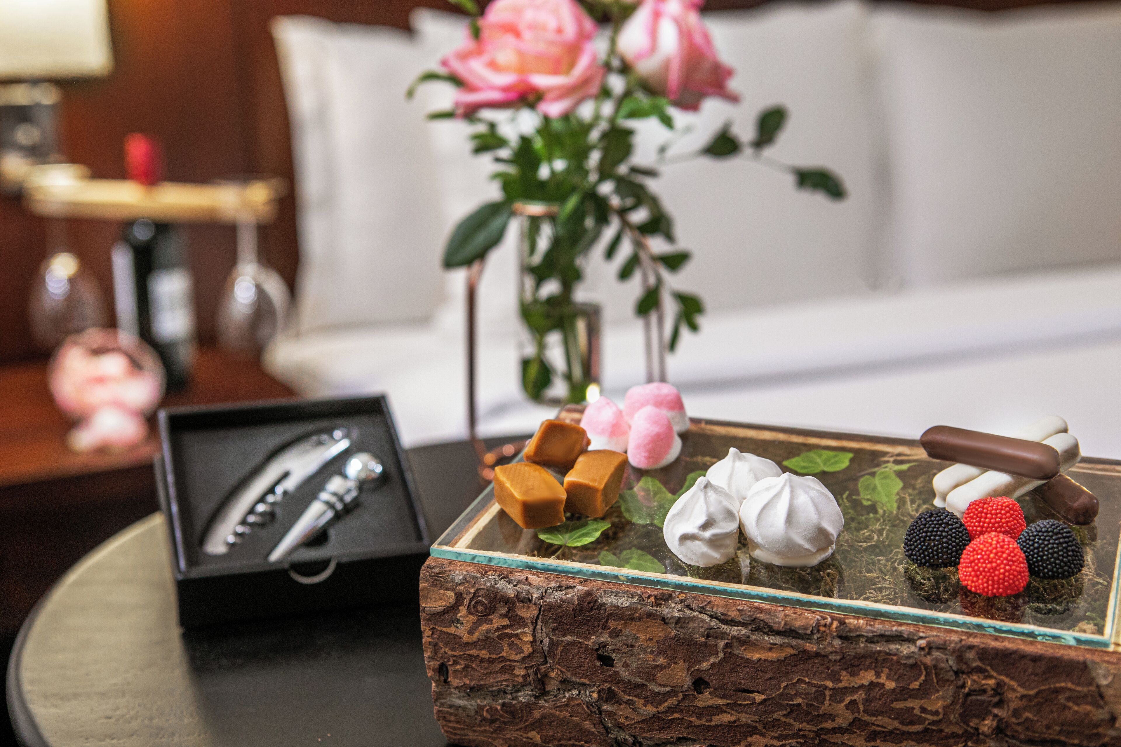 Una bandeja de postres con bombones y bayas sobre una mesa de madera rústica, una caja negra con un bolígrafo y un jarrón con rosas rosadas en un ambiente de hotel.